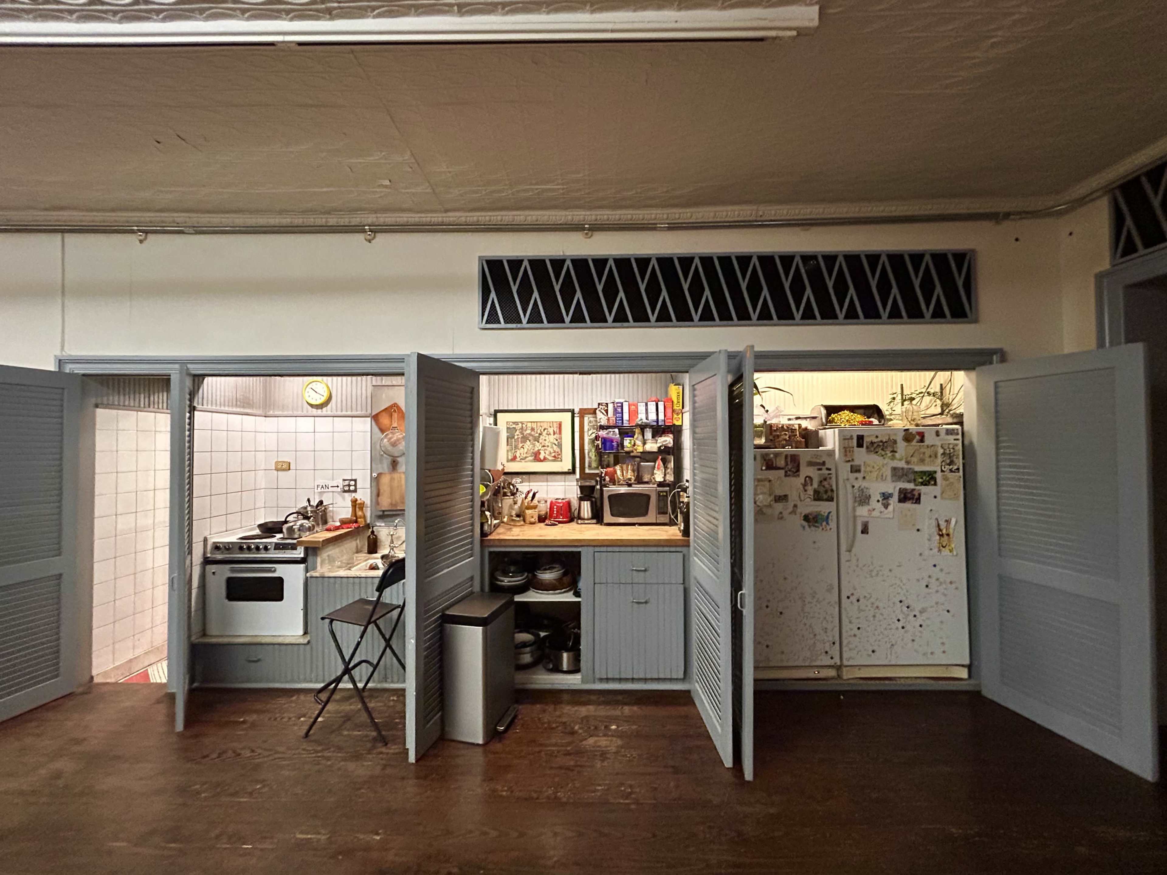 The image shows a small kitchen area with open louvered doors, featuring white appliances, a stove, a microwave, and a table with chairs, all set against wooden flooring.