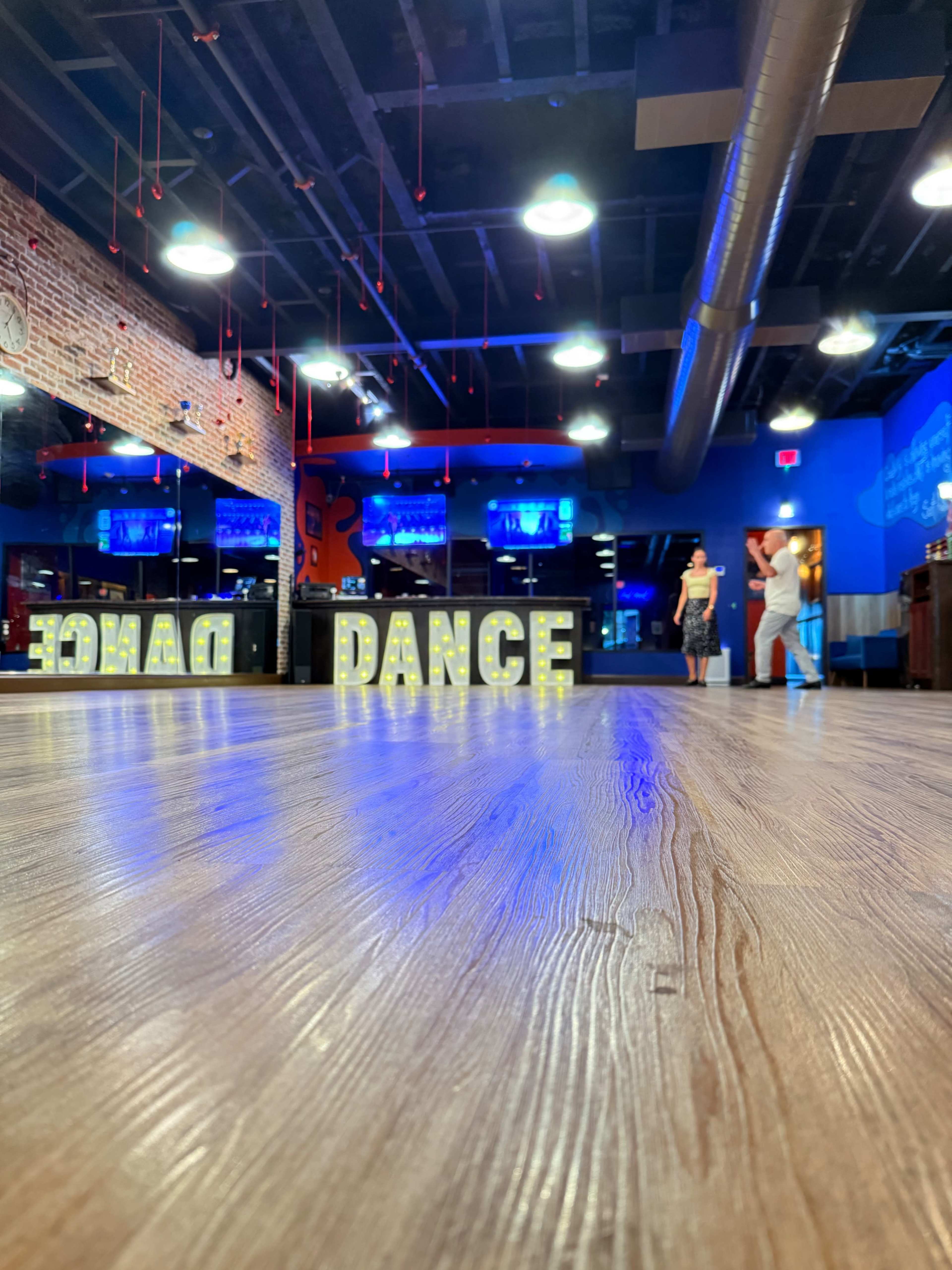The image shows an empty dance studio with illuminated letters spelling "DANCE" on a wooden floor, and two people are seen in the background.