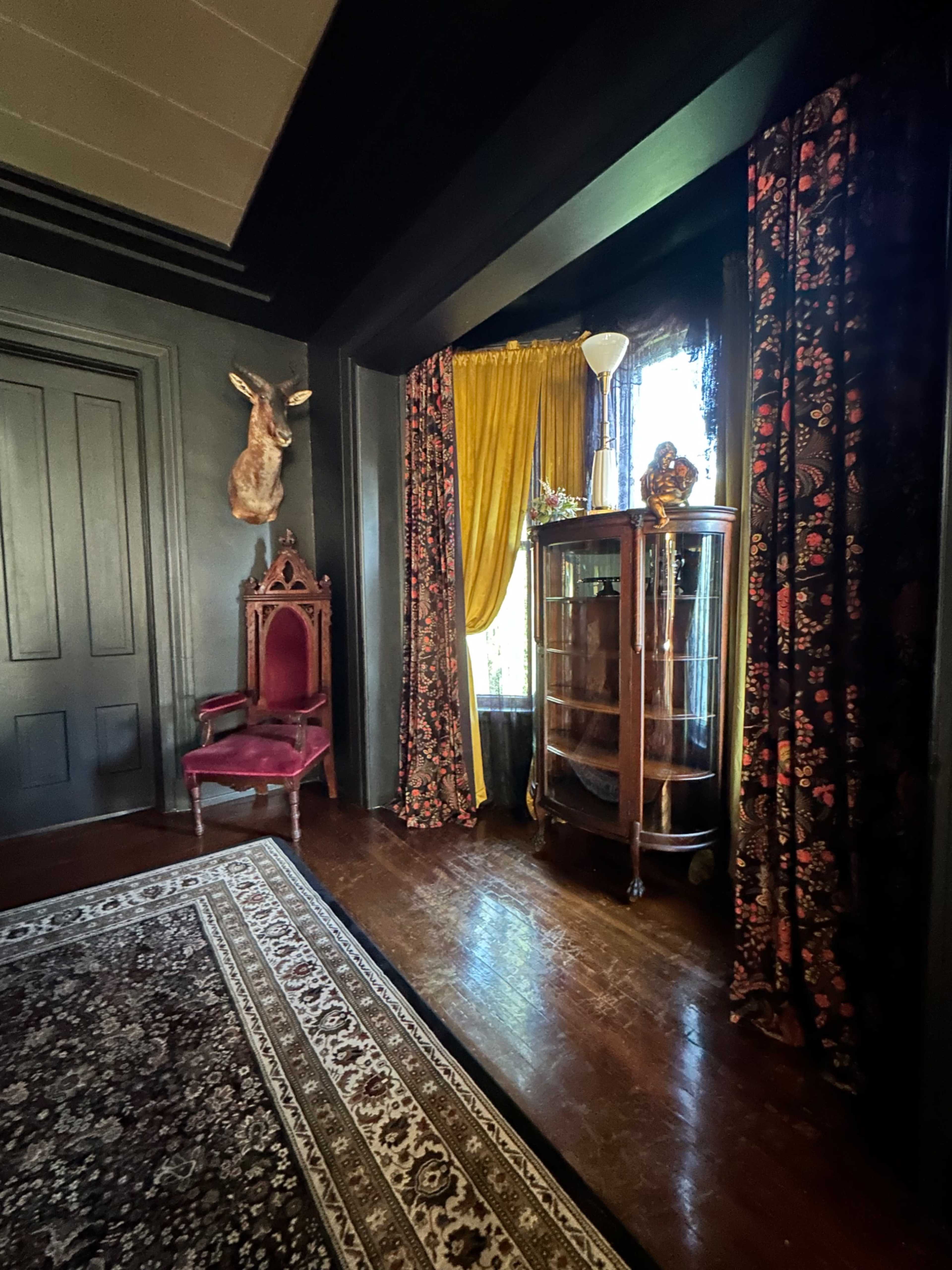 The room features dark walls, a patterned rug, a red chair, a glass display cabinet, and large windows with golden drapes.