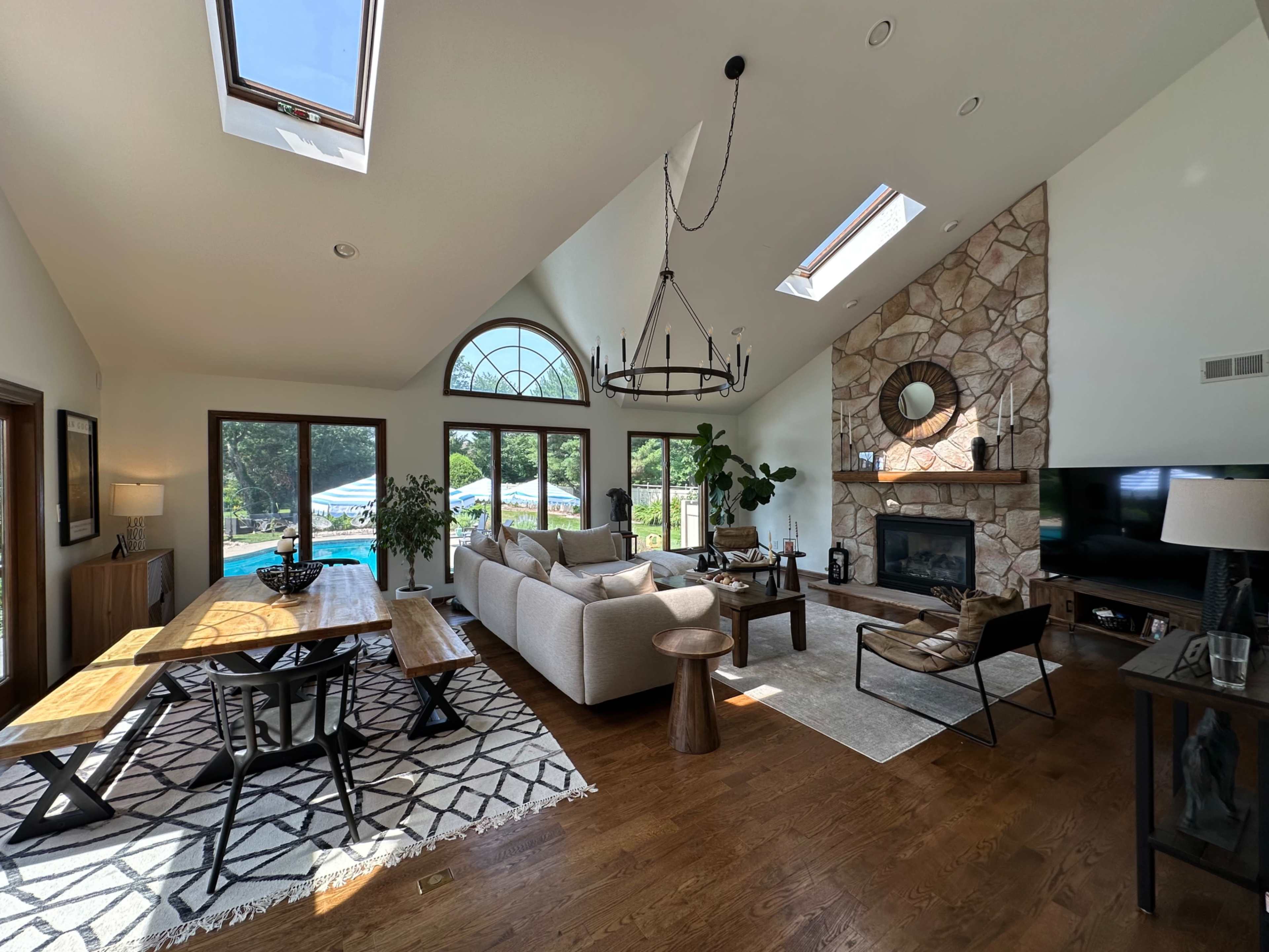 The image shows a modern living space with a wooden dining table, a large sectional sofa, and a stone fireplace, all surrounded by large windows that overlook a pool area.
