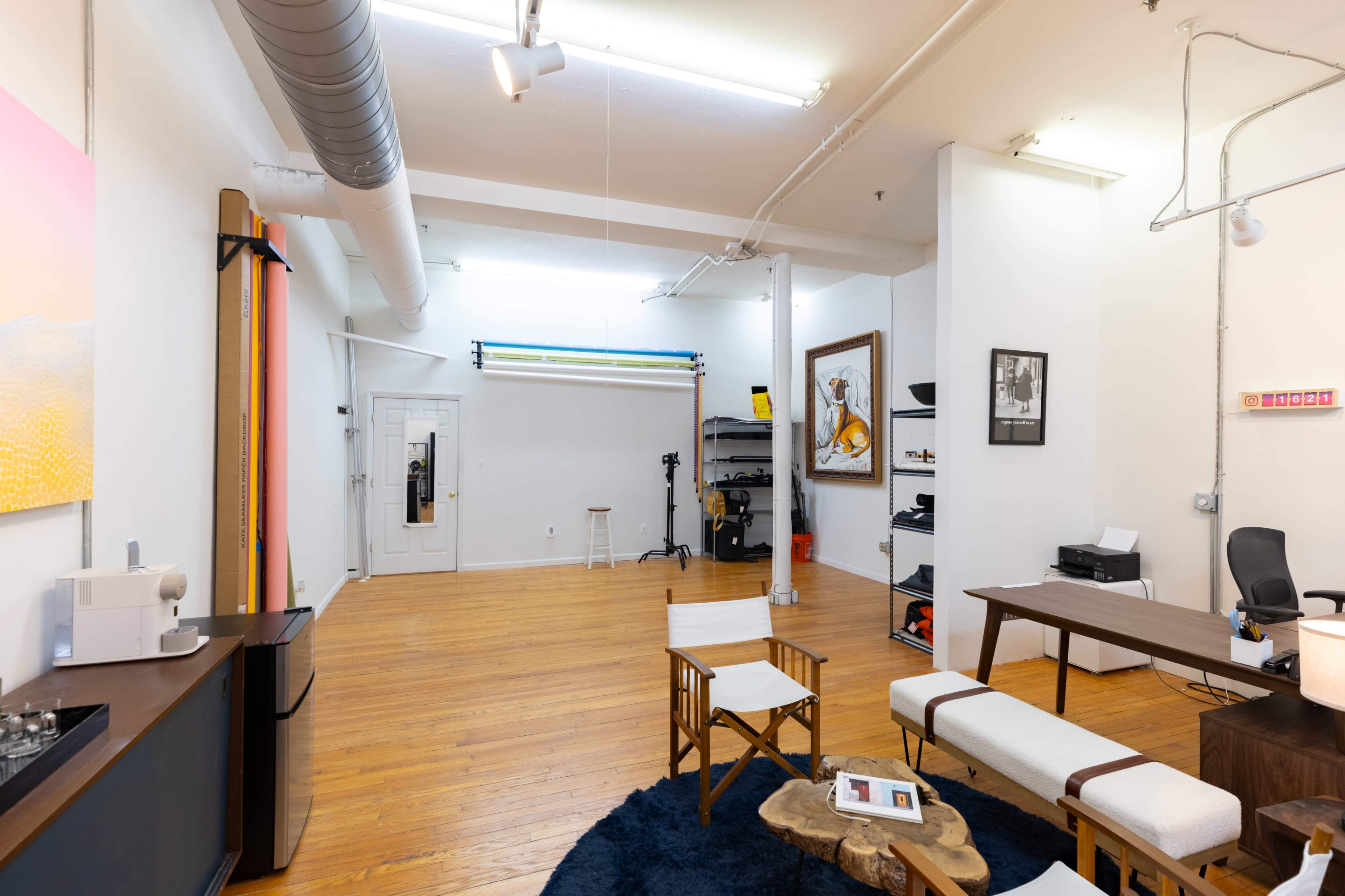 The image shows a spacious, well-lit studio interior featuring wooden flooring, minimalist furniture, and various equipment along the walls.