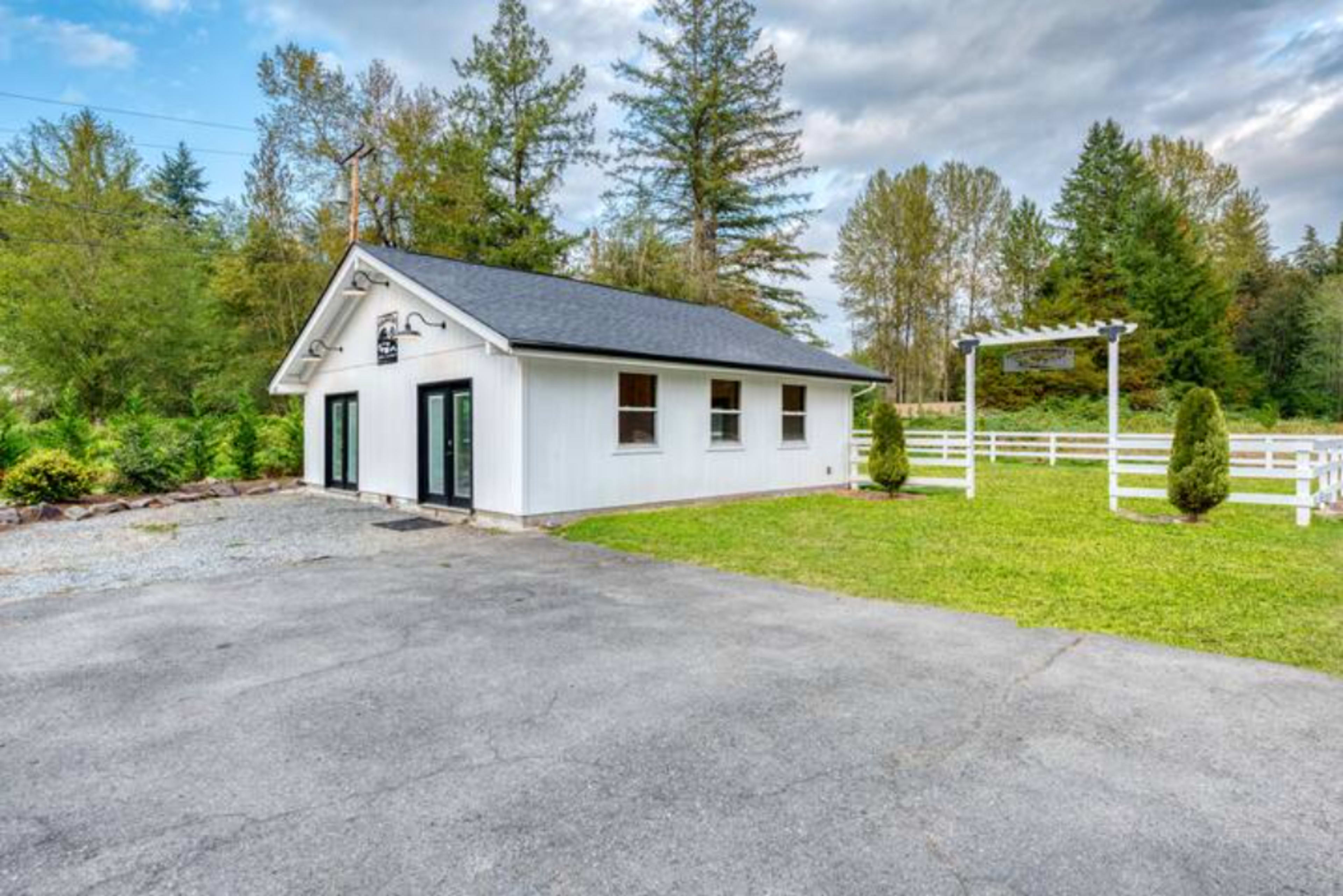 A small white building with large windows, set on a gravel driveway next to a green lawn and surrounded by trees, with a white fence visible in the background.