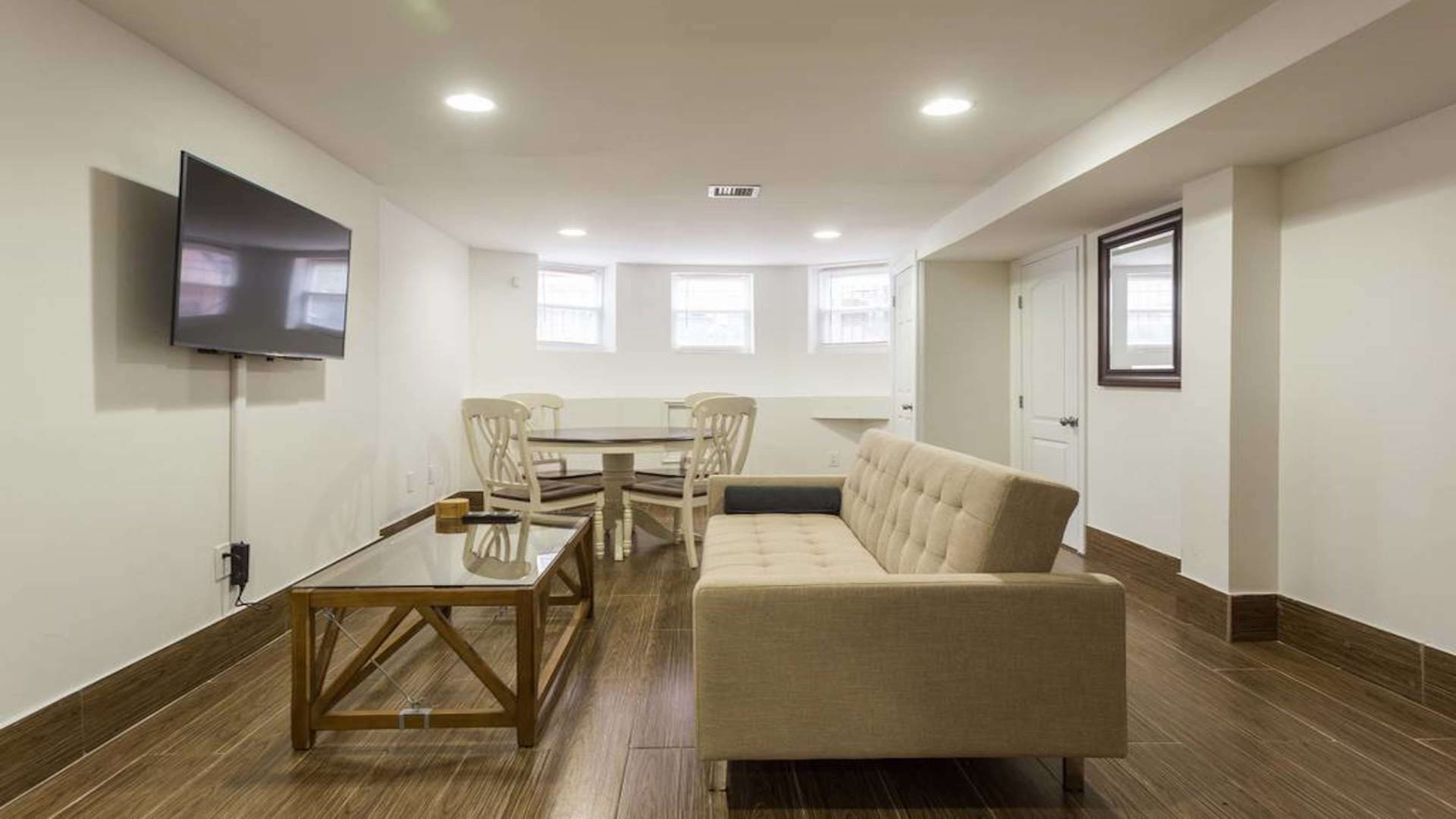 The image shows a basement living area featuring a beige sofa, a glass coffee table, and a dining table with chairs, all surrounded by white walls and bright lighting.
