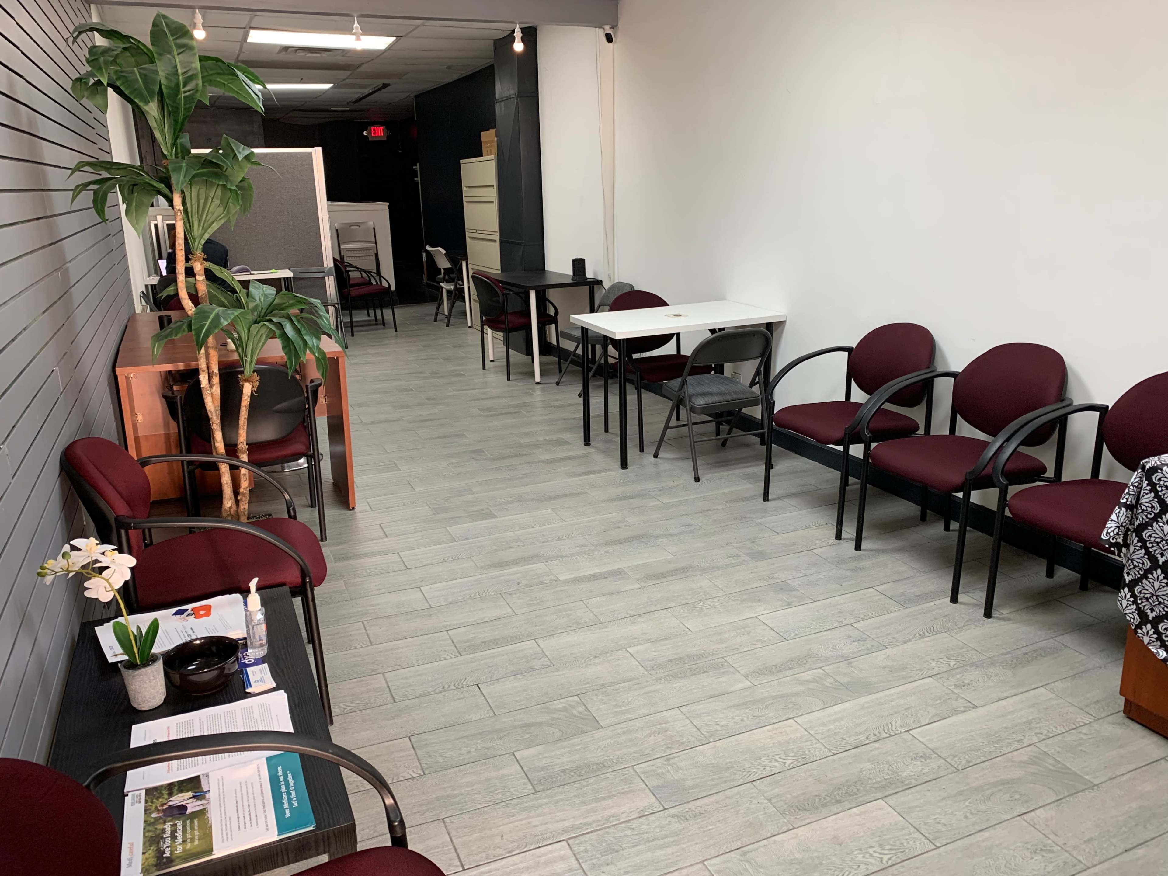 A waiting area with several maroon chairs, a potted plant, and a table, set against a light-colored wall and gray tile floor.