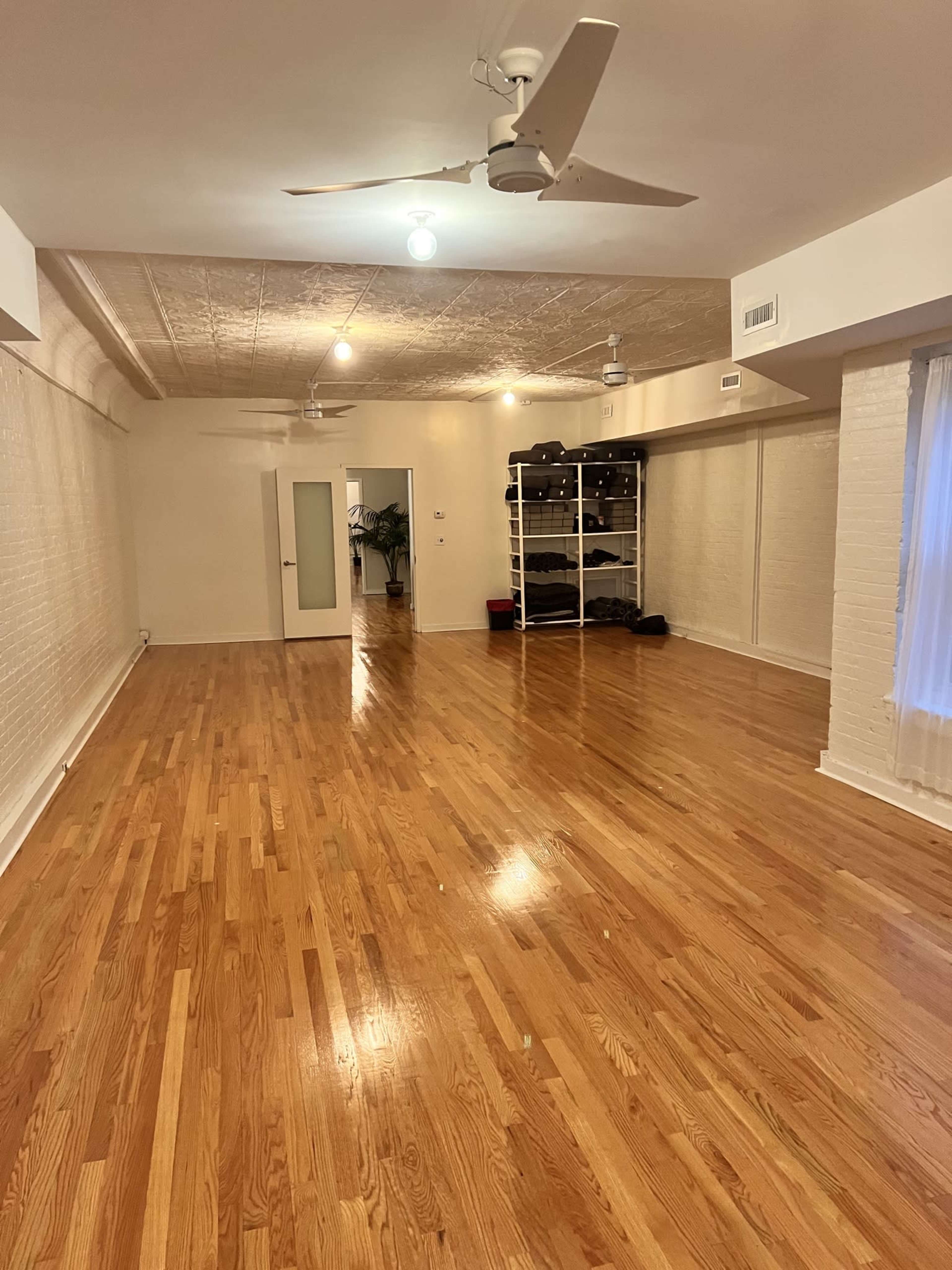 A large, empty room with hardwood flooring, white walls, and multiple ceiling fans, leading to an adjoining room with a shelving unit.
