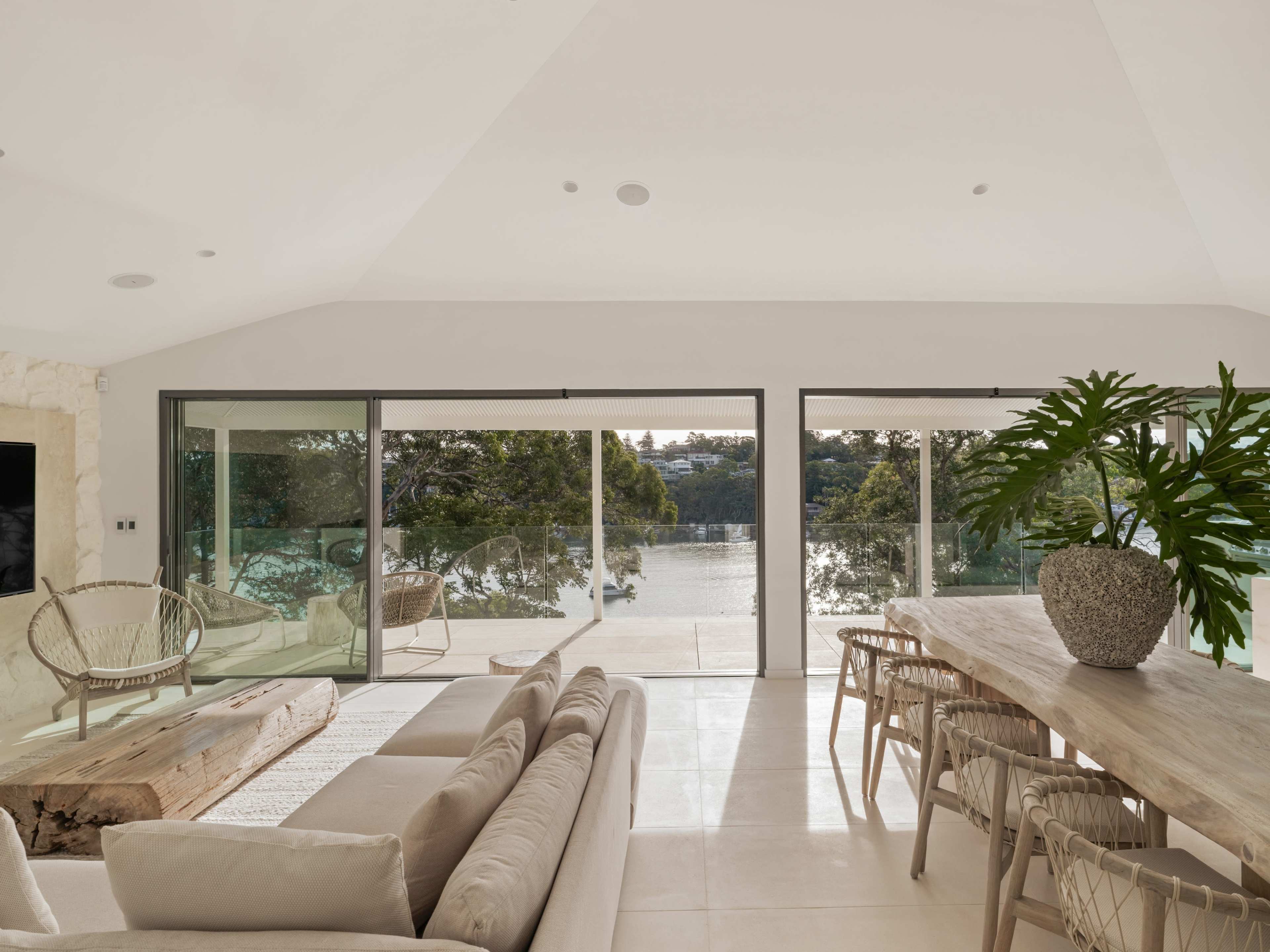 The image shows a modern living space with large glass doors that open to a waterfront view, featuring a beige sofa, a wooden dining table, and a large potted plant.