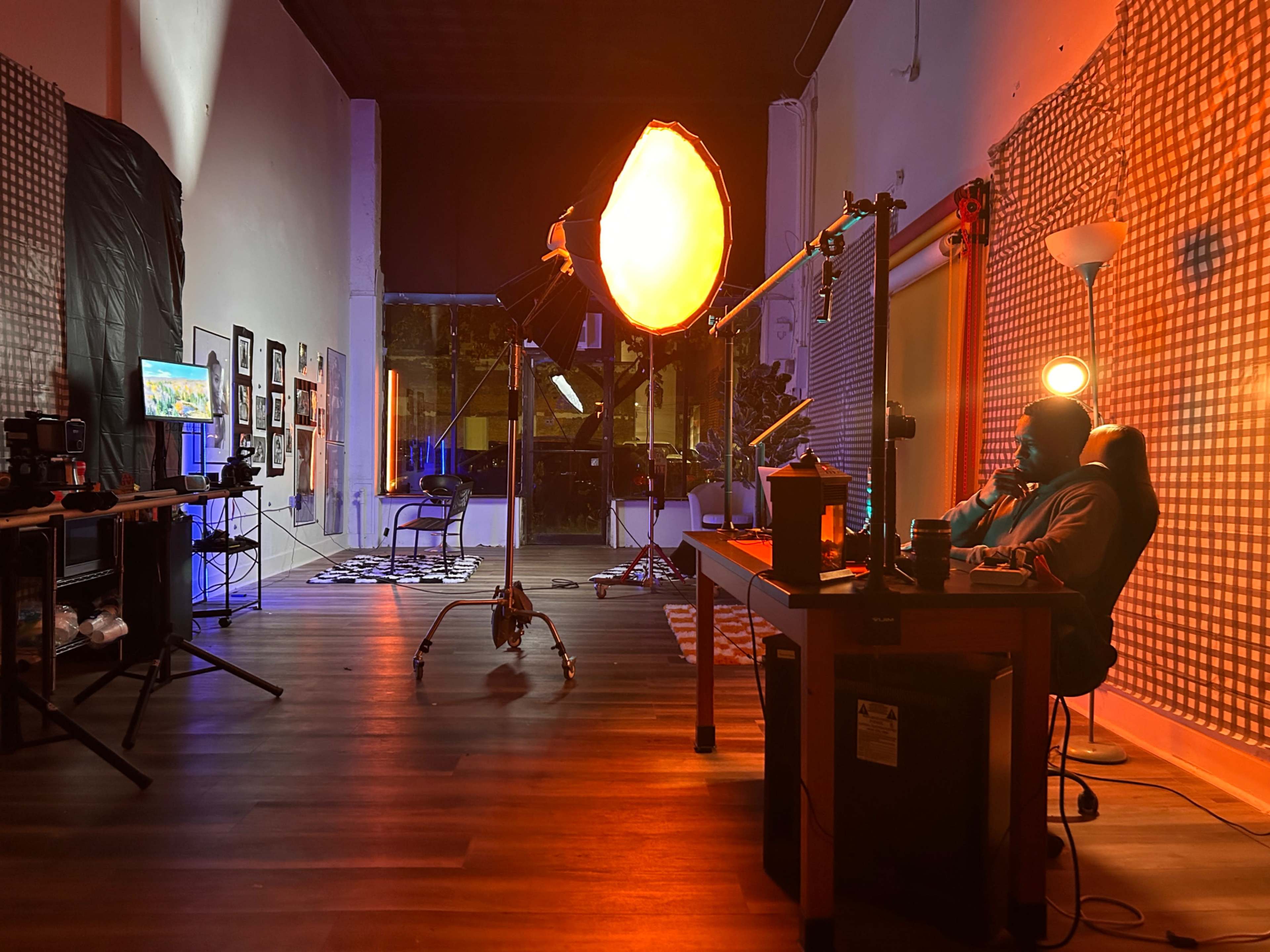 A dimly lit studio features a person sitting at a desk while illuminated by a large softbox light, surrounded by photography equipment and framed photos on the wall.