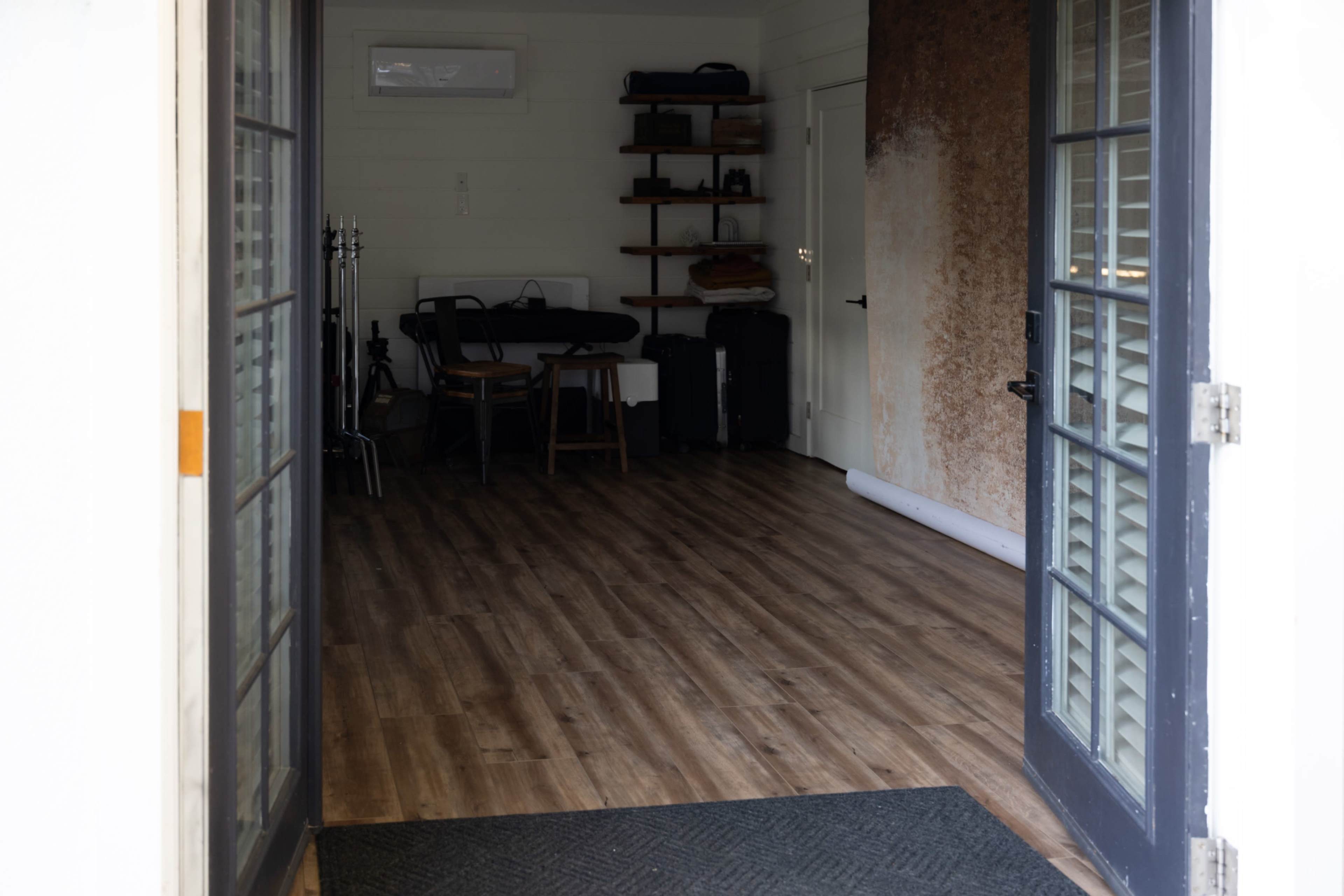 The image shows an interior space viewed through a set of double doors, featuring wooden flooring, a table with chairs, and shelves against a wall.
