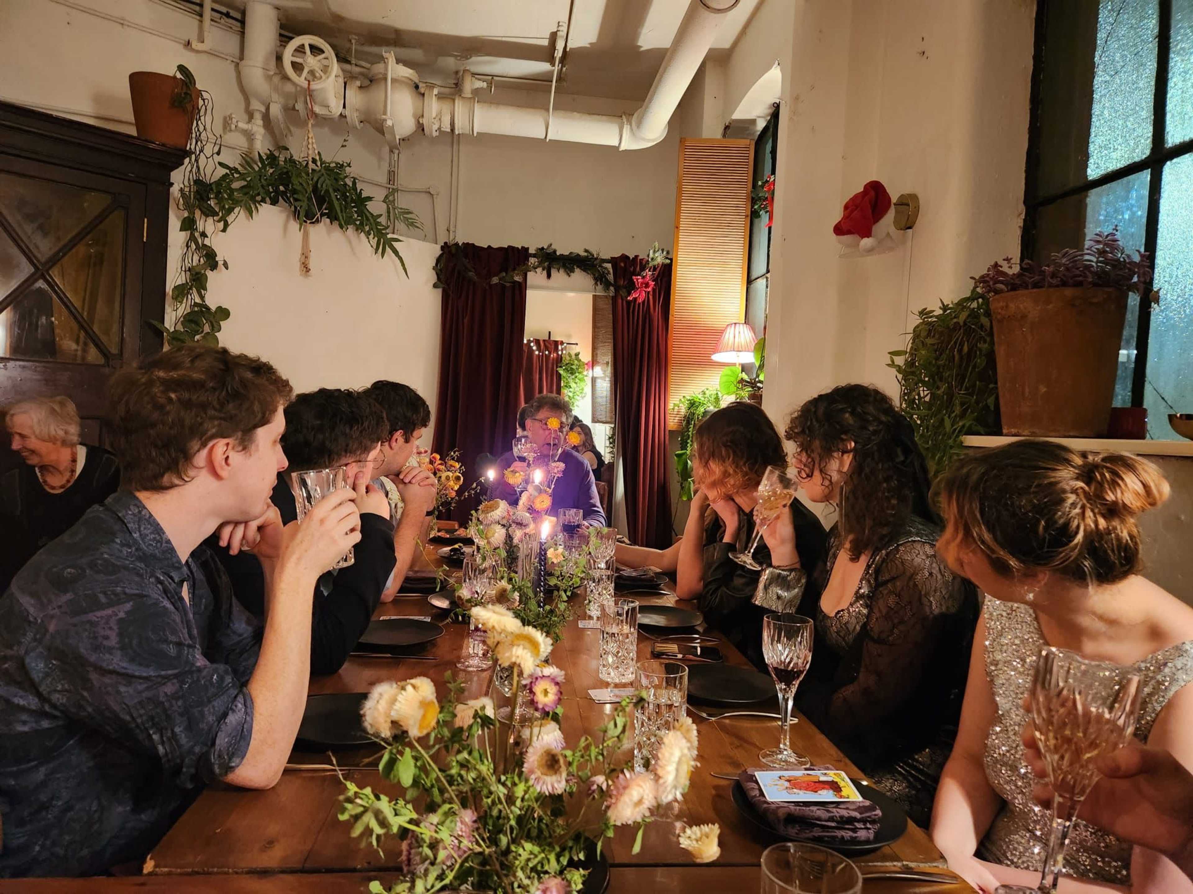 A group of people sits around a wooden dining table adorned with flowers and candles in a warmly lit room.