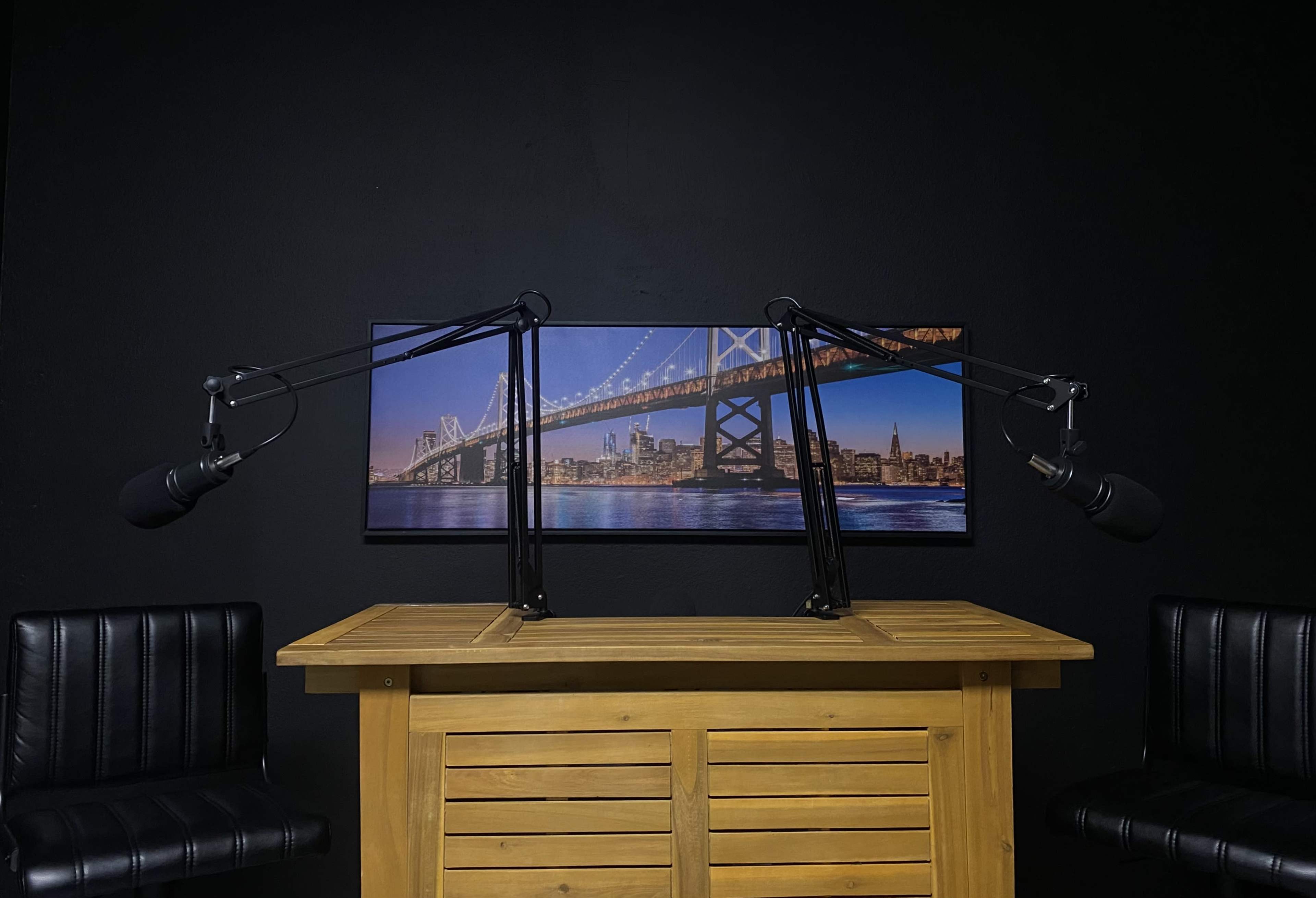 A podcast studio features a wooden table with two adjustable microphone mounts and a backdrop of a bridge at night.
