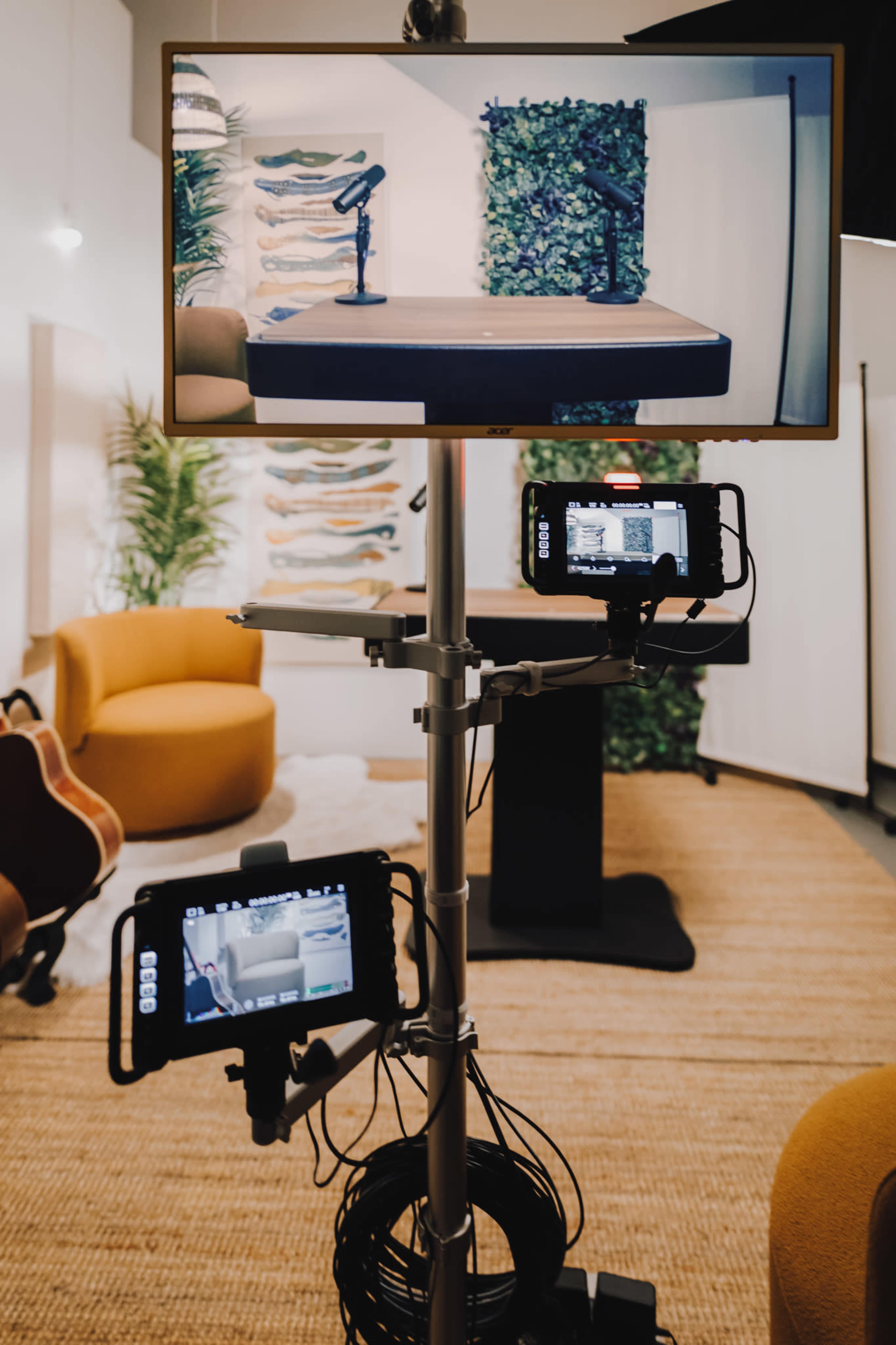A mounted screen displays a table with microphones in a studio setting, featuring plants and cozy seating in the background.