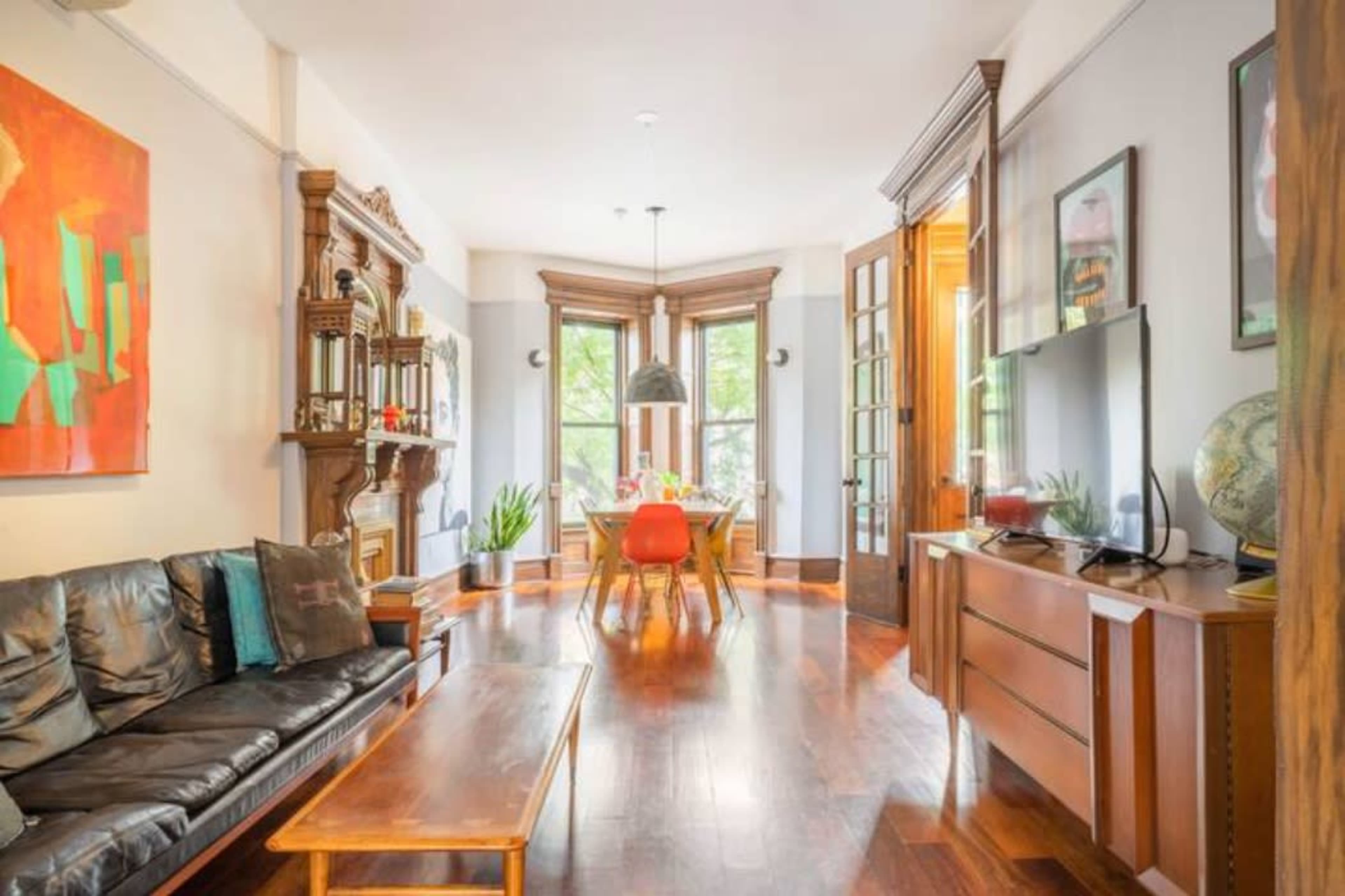 The image shows a living room with a dark leather sofa, a wooden coffee table, and a dining area featuring a large window and a circular table surrounded by chairs.