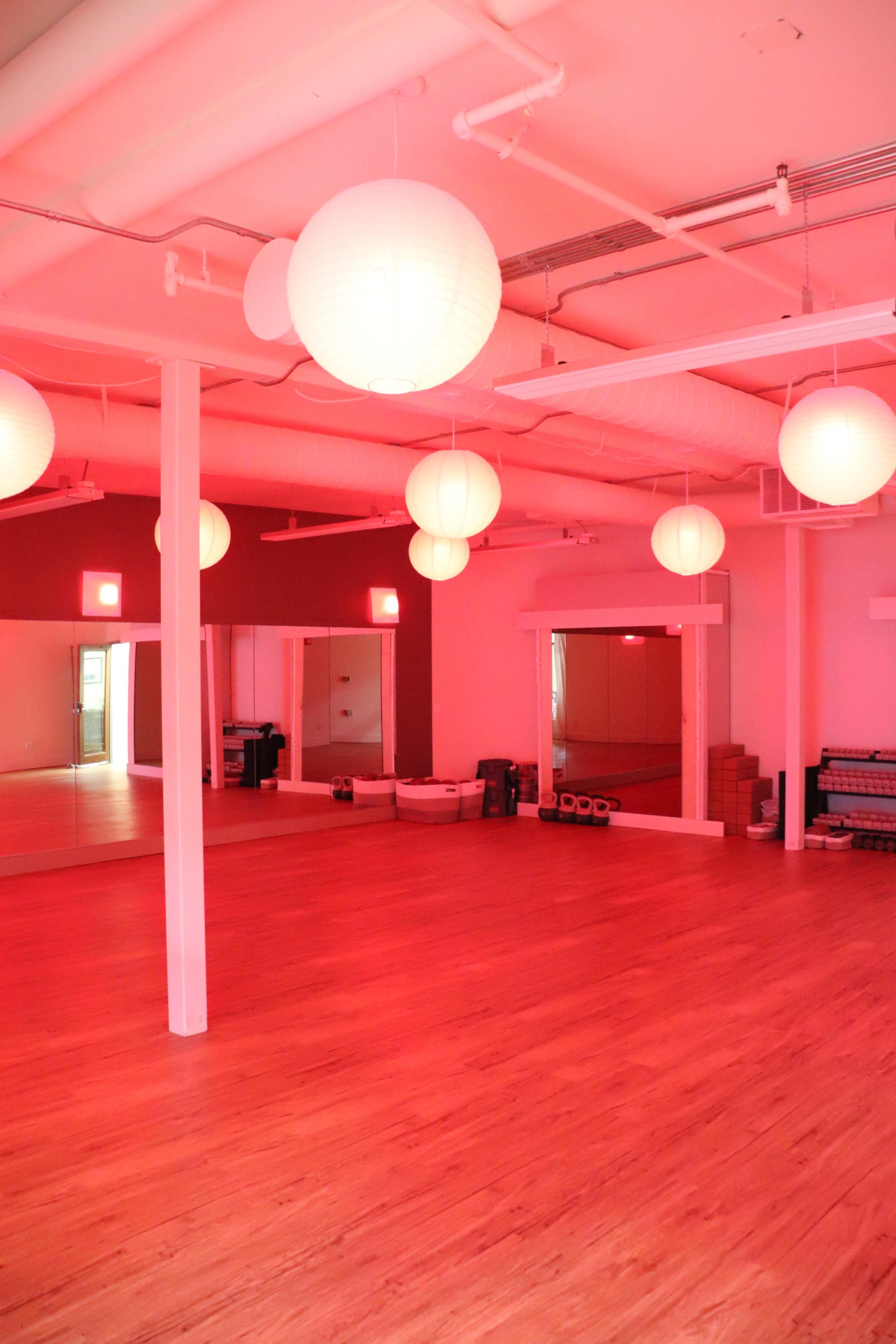 The image shows a brightly lit studio with wooden floors, paper lanterns hanging from the ceiling, and mirrors on one wall.