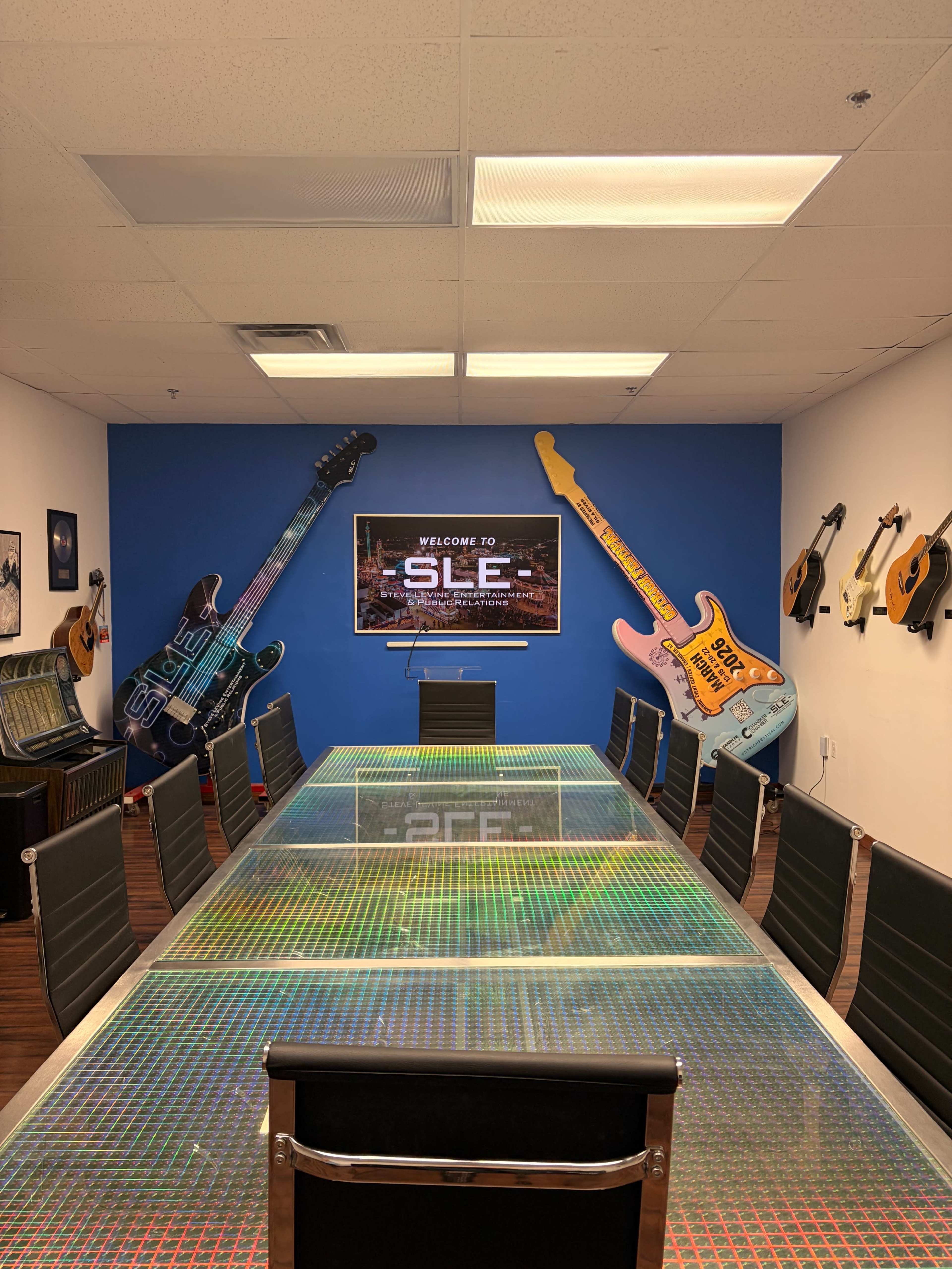A conference room features a long glass table surrounded by black chairs, with guitars displayed on the walls and a screen showcasing the text "WELCOME TO SLE."