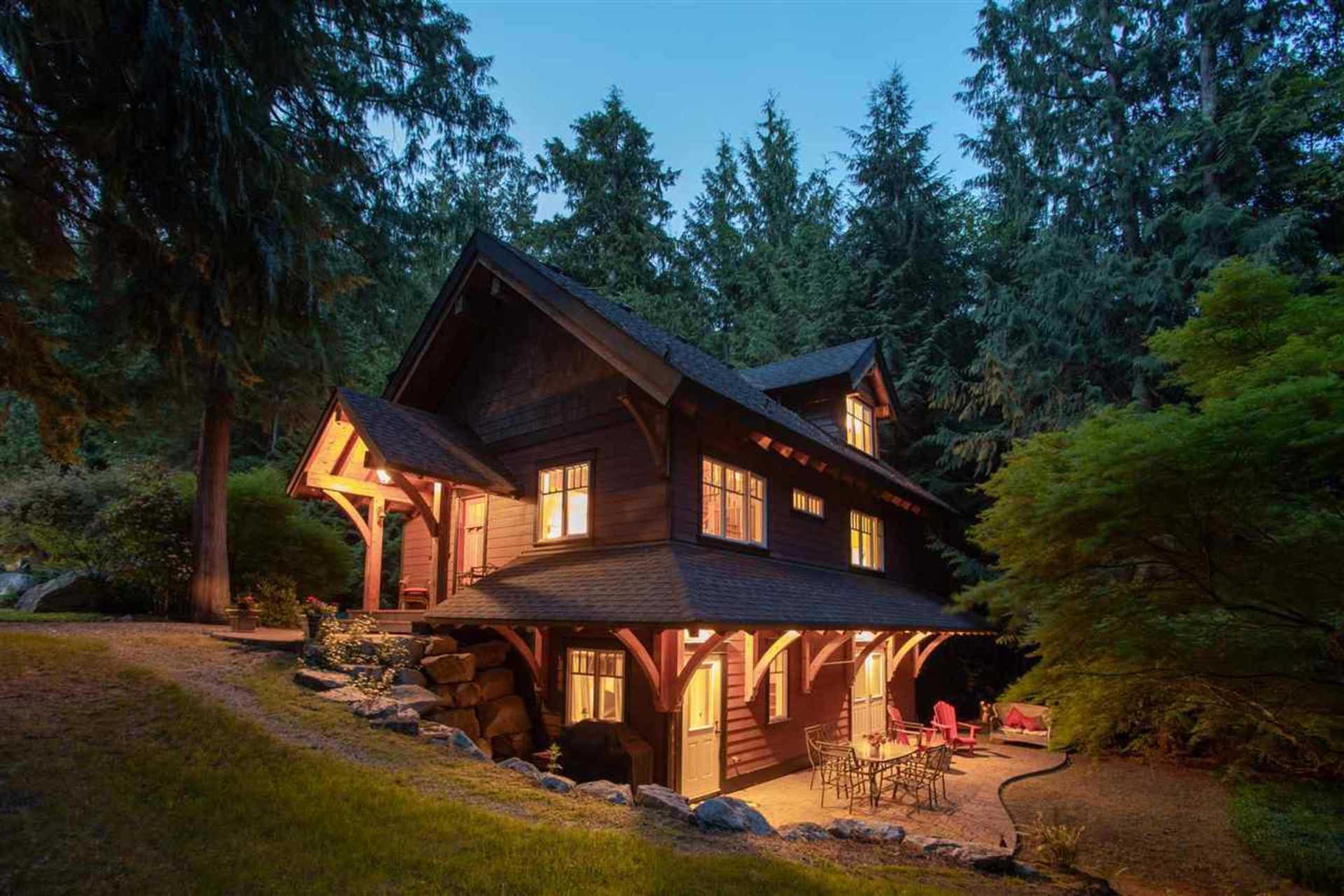 A wooden house sits amidst tall trees, illuminated by lights from its windows, with seating arranged outside.