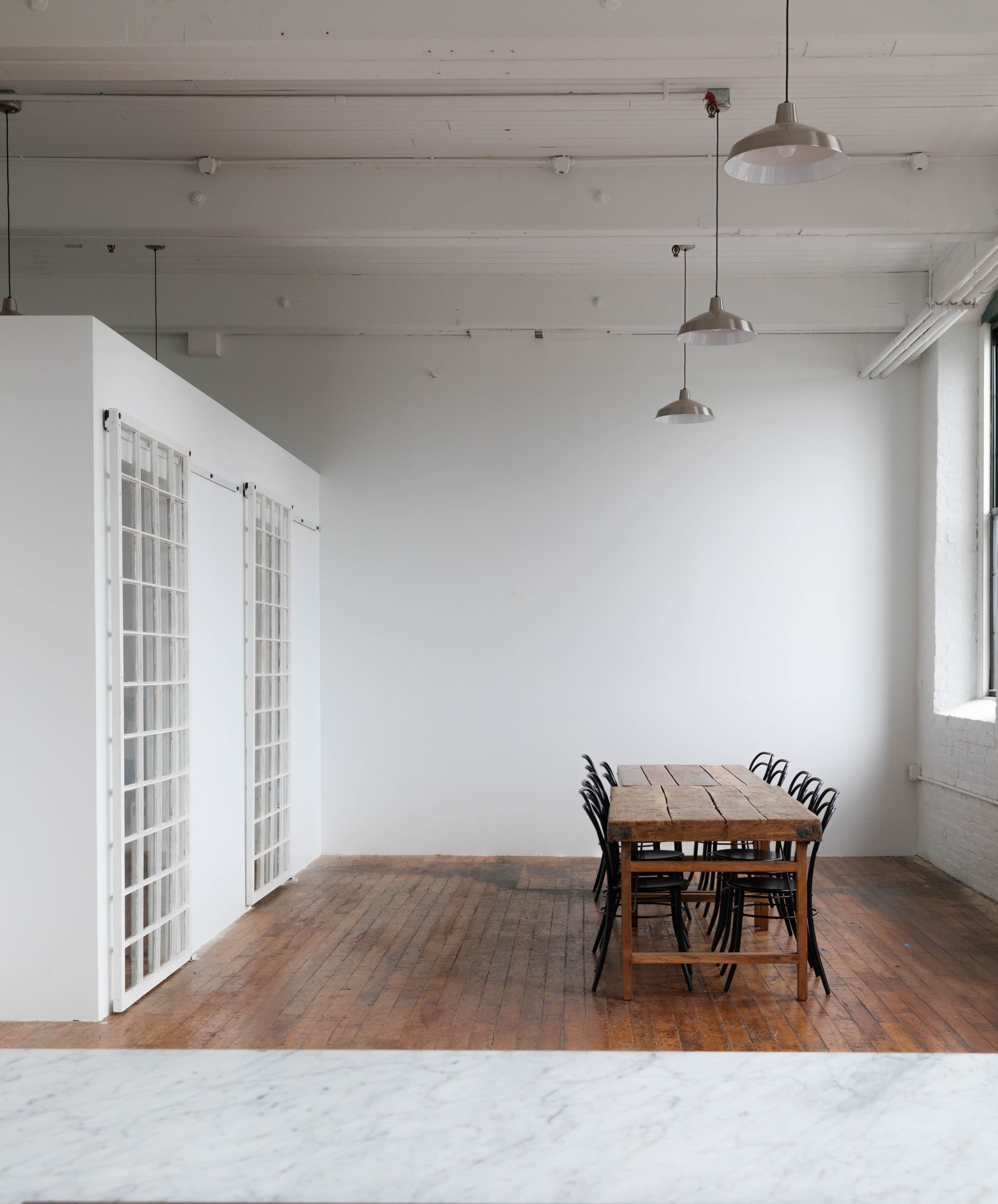 A spacious, minimalist room features a long wooden table with black chairs, facing a white wall and large windows.