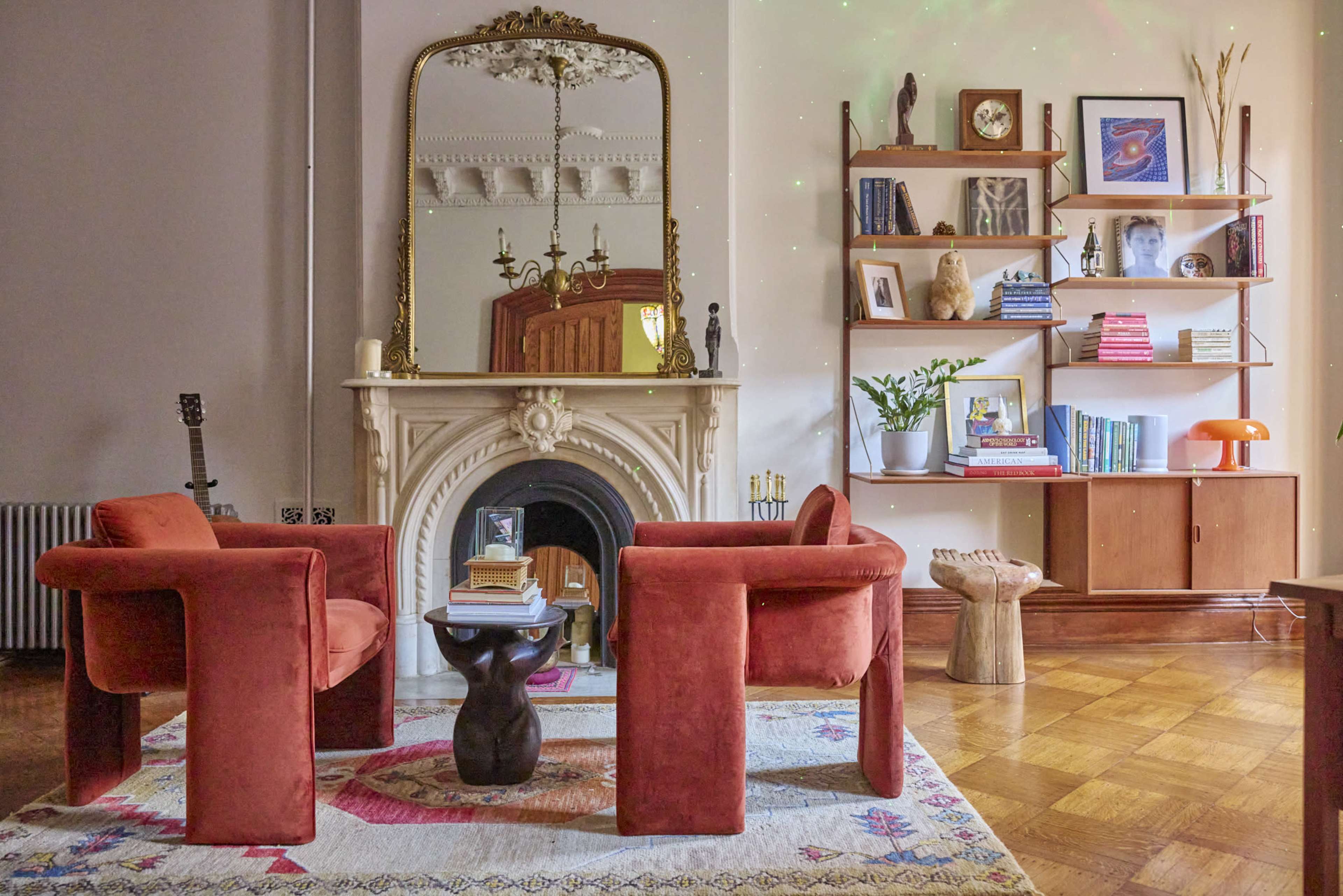 A cozy living room features two red armchairs facing a small black table, with a marble fireplace and a shelf filled with books and decorative items in the background.