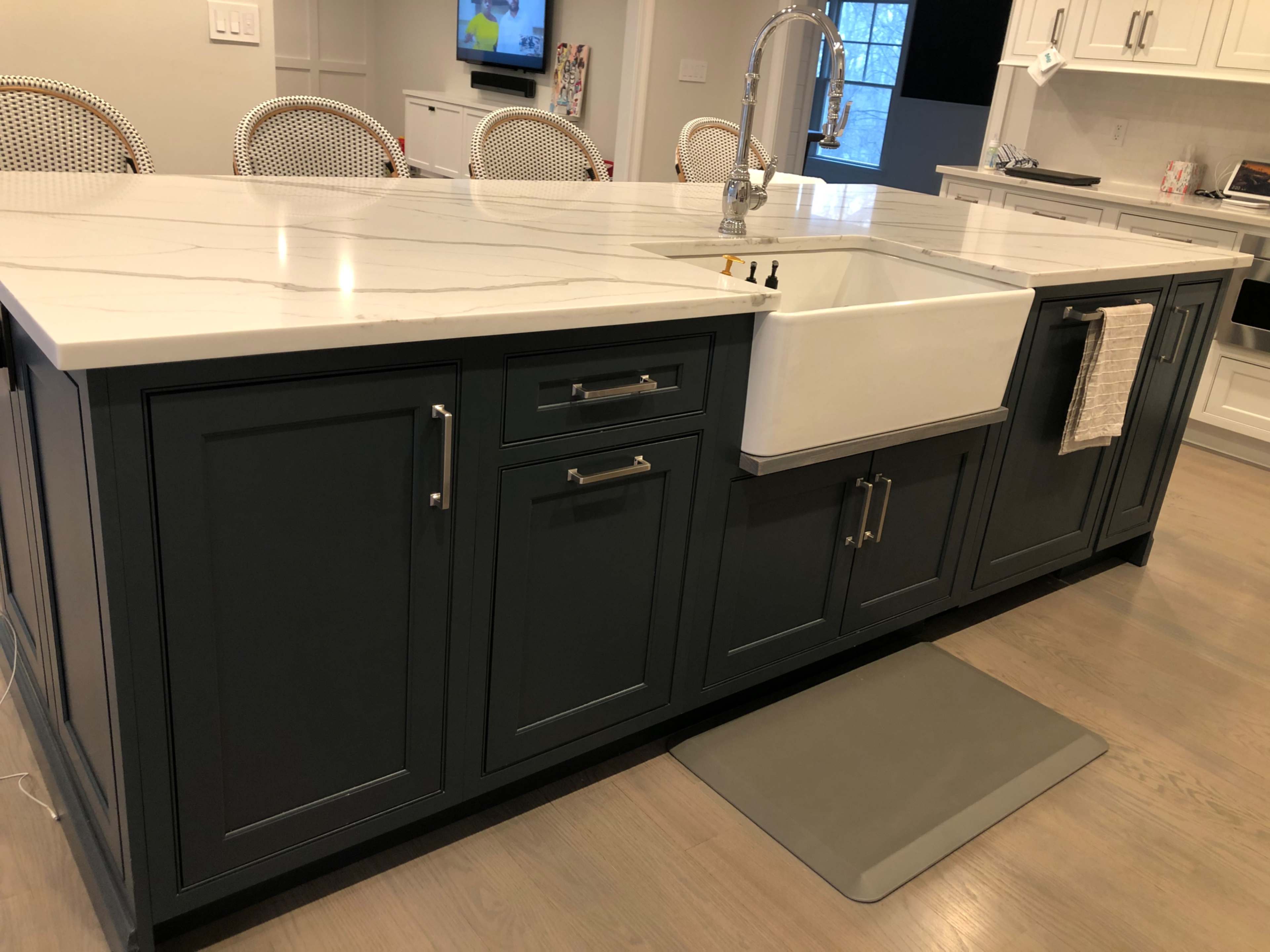 A large kitchen island with a white marble countertop and a farmhouse sink is shown, surrounded by modern cabinetry and a mat on the floor.