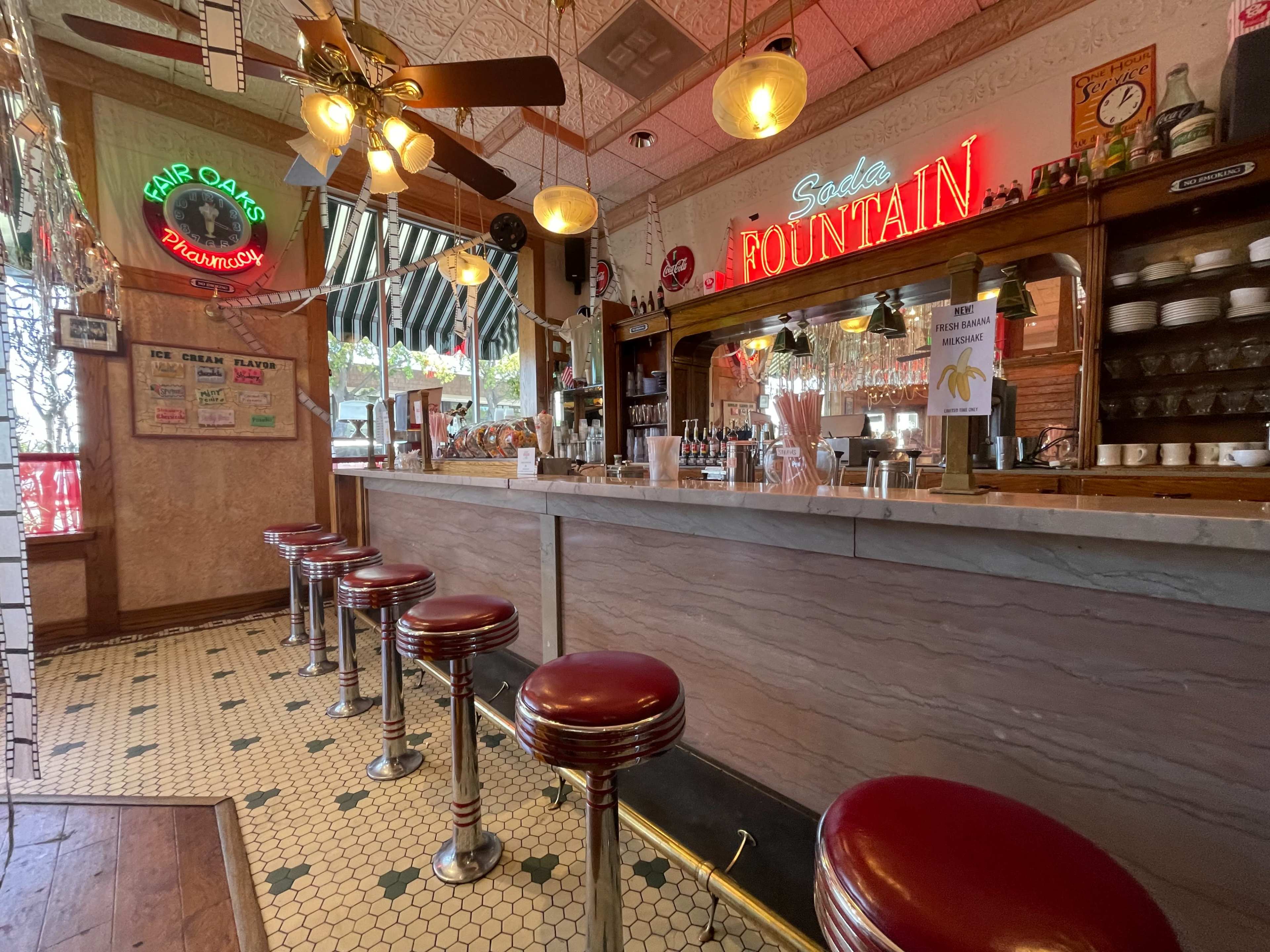 A vintage soda fountain with red barstools, a marble counter, and retro signage in a well-lit diner setting.
