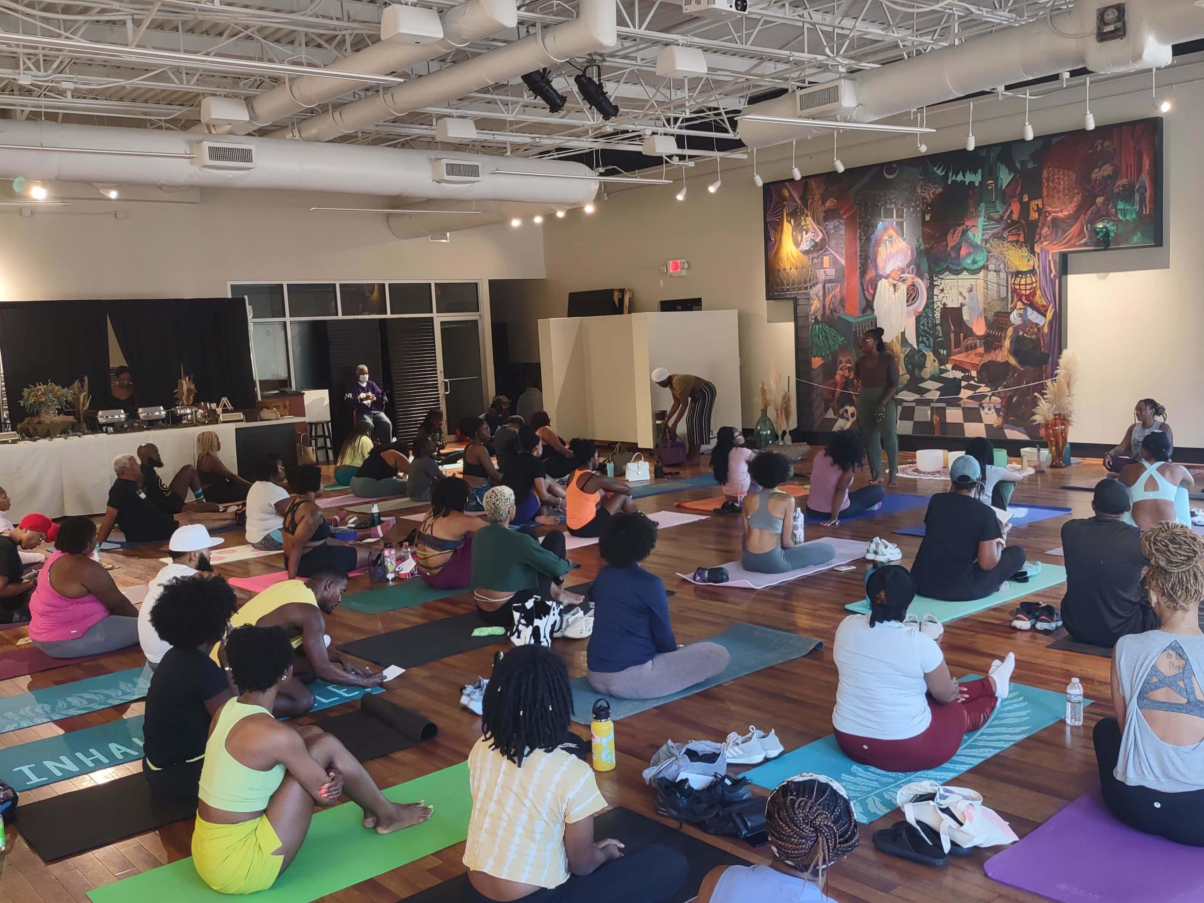 A large group of individuals participates in a yoga class inside a studio featuring a colorful mural.