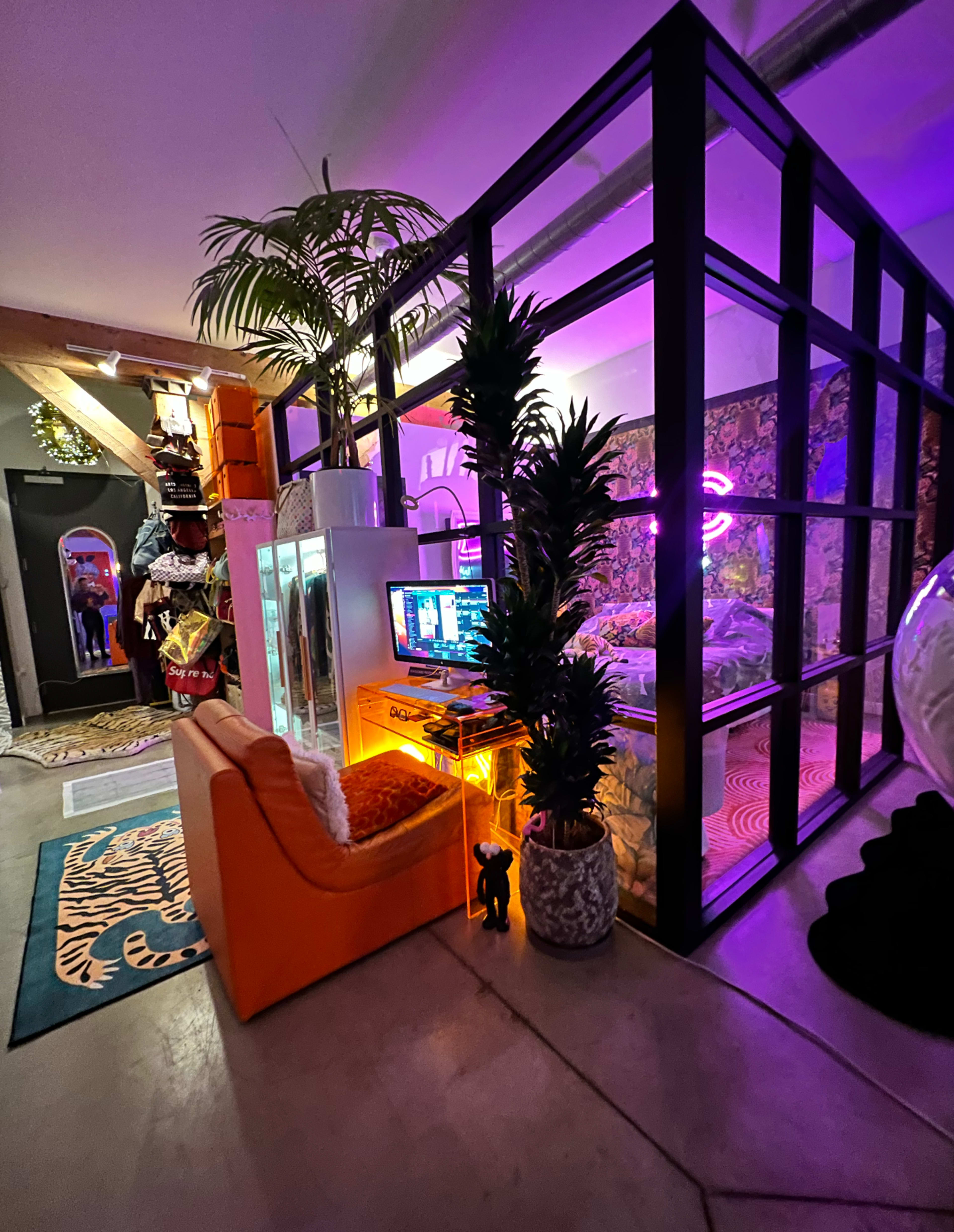 The image shows a modern, stylish room featuring a glass partition, eclectic decor, a gaming setup with neon lights, an orange sofa, and potted plants.
