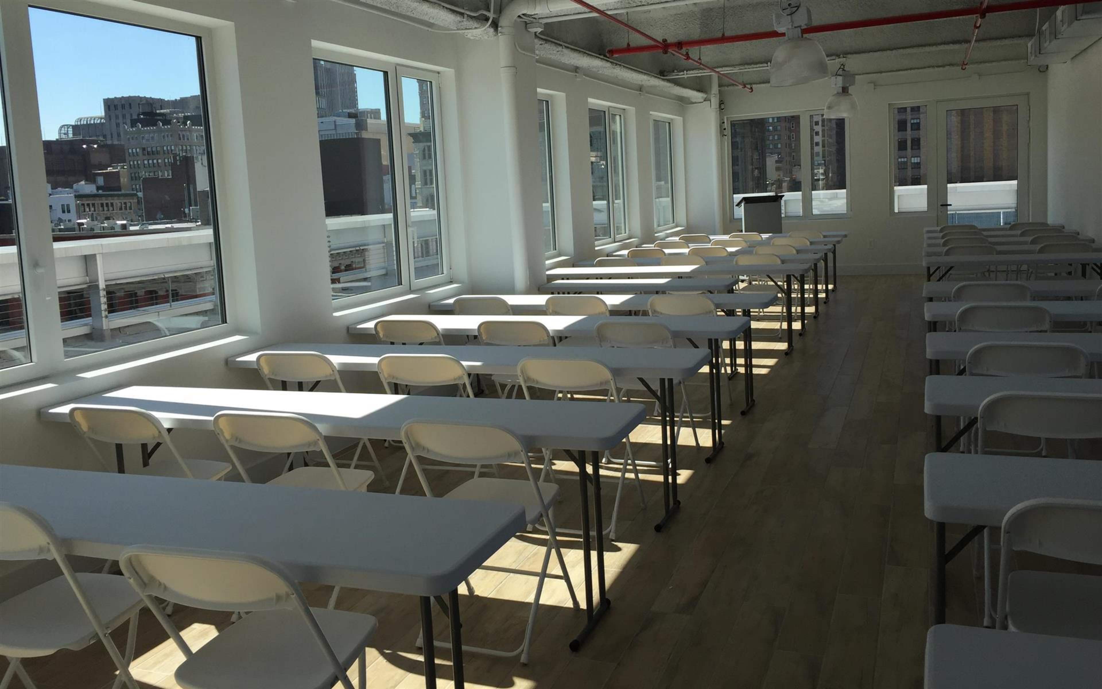 A bright classroom with rows of white tables and chairs arranged next to large windows.