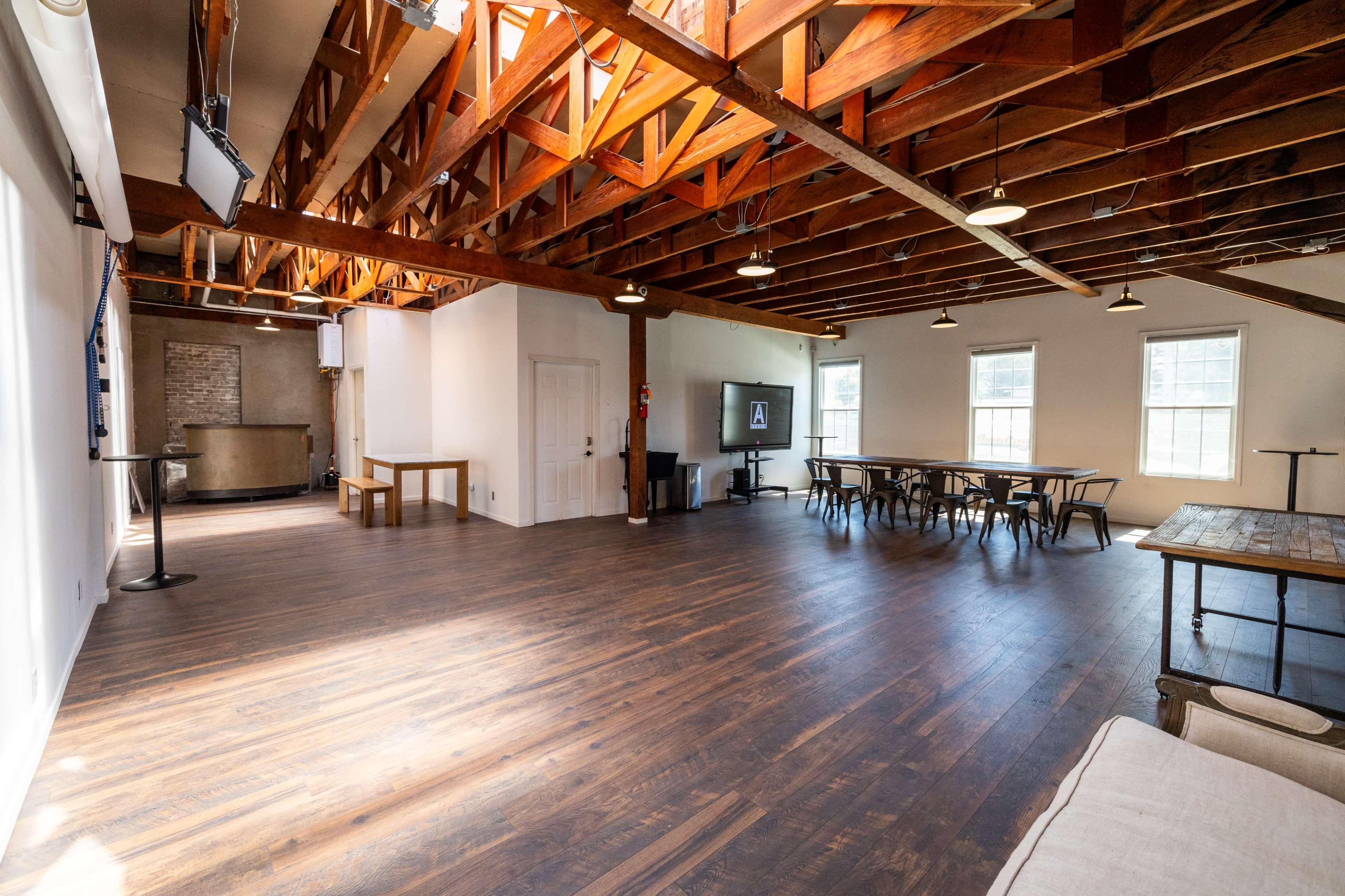 A spacious room with wooden beams, large windows, and various seating arrangements, including tables and chairs.