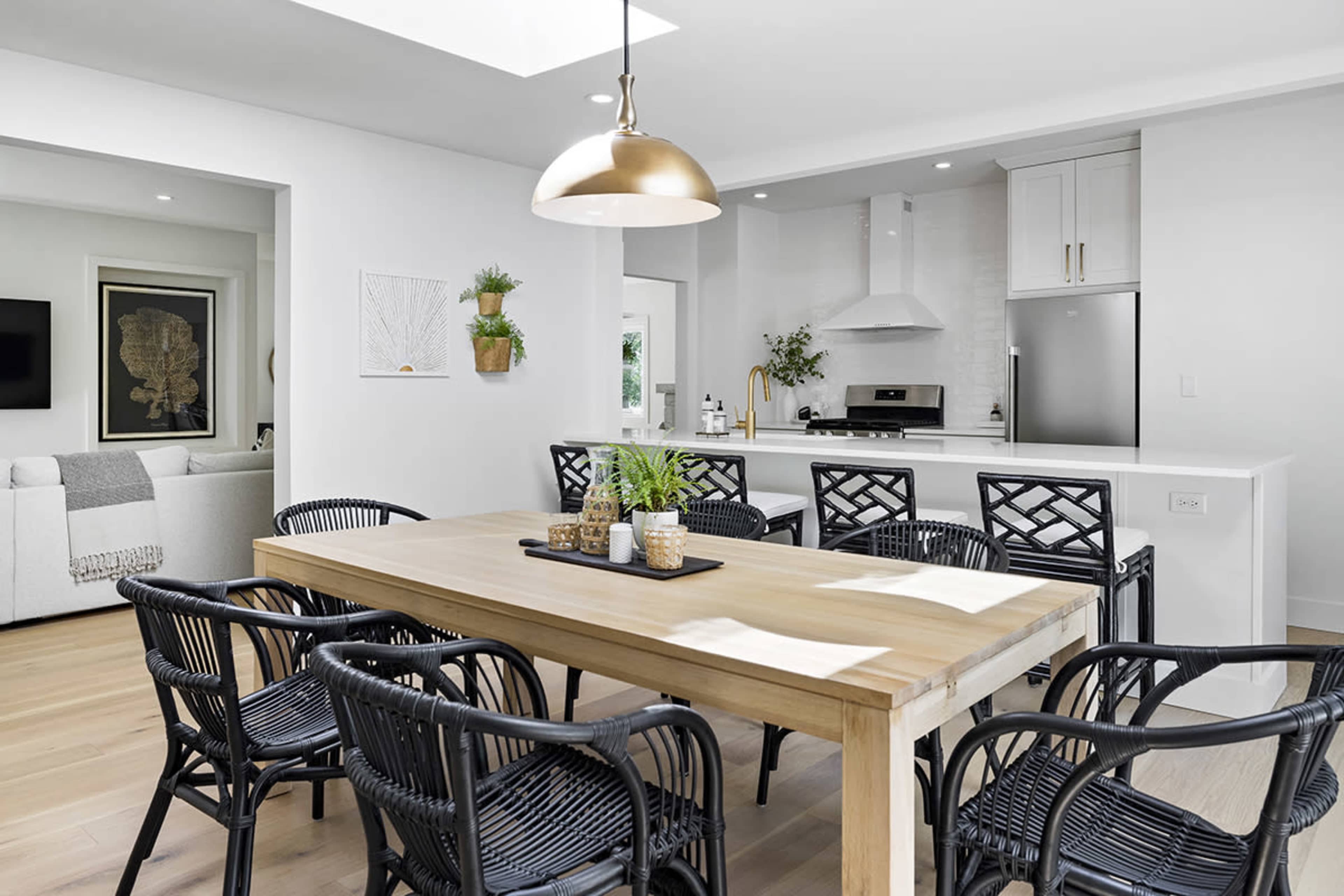 A spacious dining area features a wooden table surrounded by black woven chairs, adjacent to a modern kitchen with white cabinetry and stainless steel appliances.