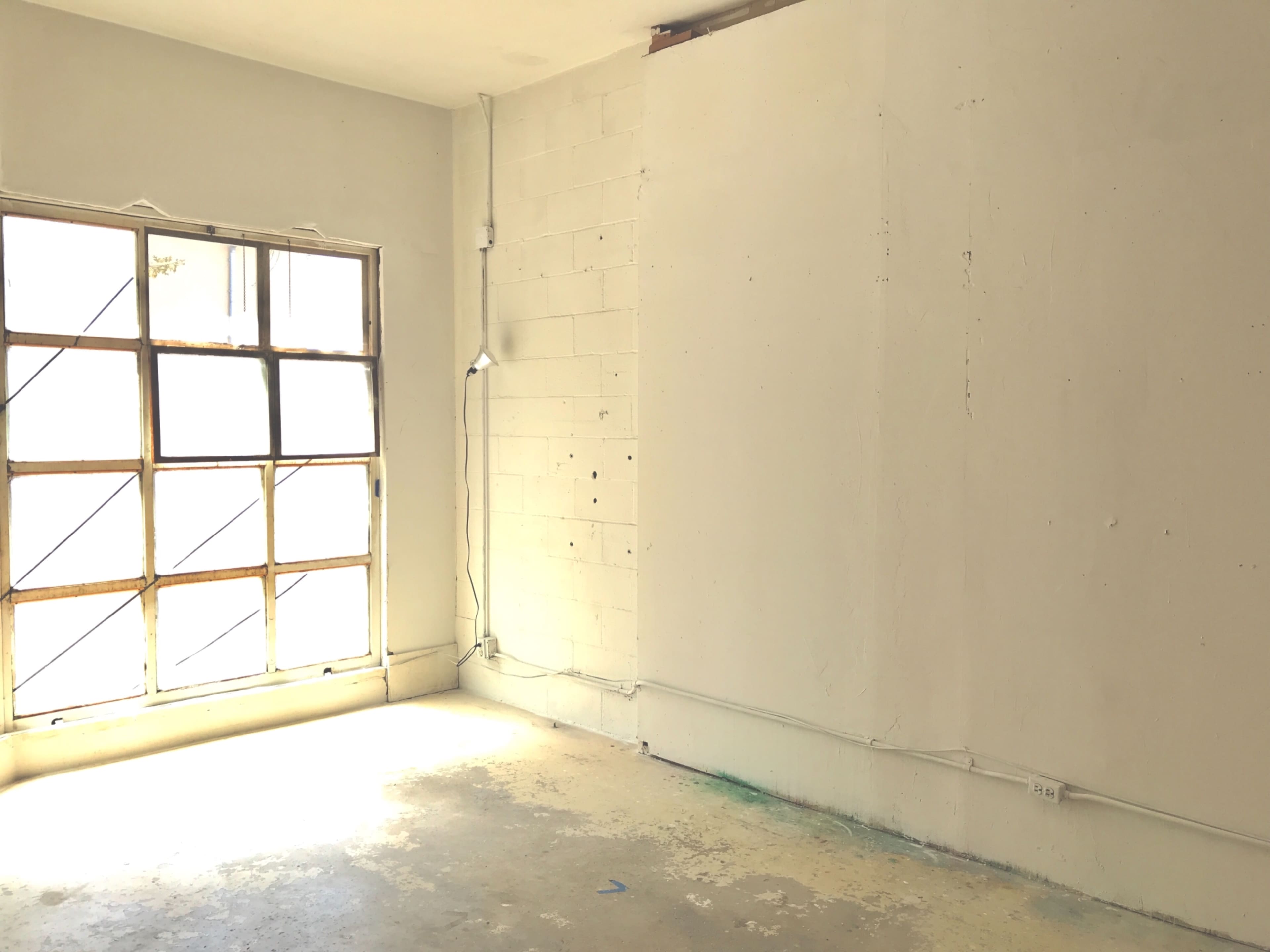 The image shows a sparsely furnished room with a large window on one side and bare walls devoid of decorations.