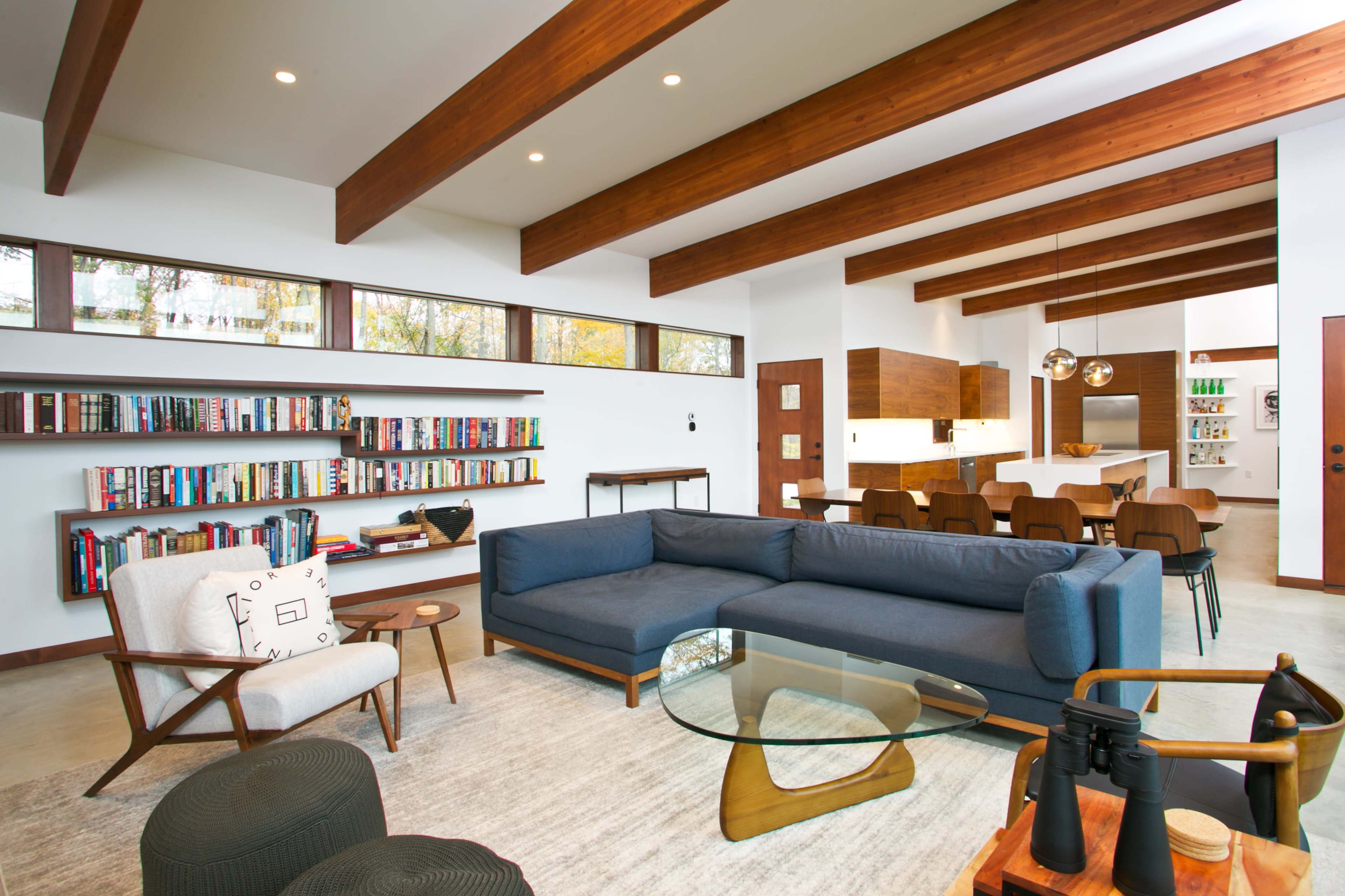 A modern living room features a large blue sectional sofa, a glass coffee table, and a bookshelf filled with various books, with wooden beams overhead and an adjoining dining area visible in the background.