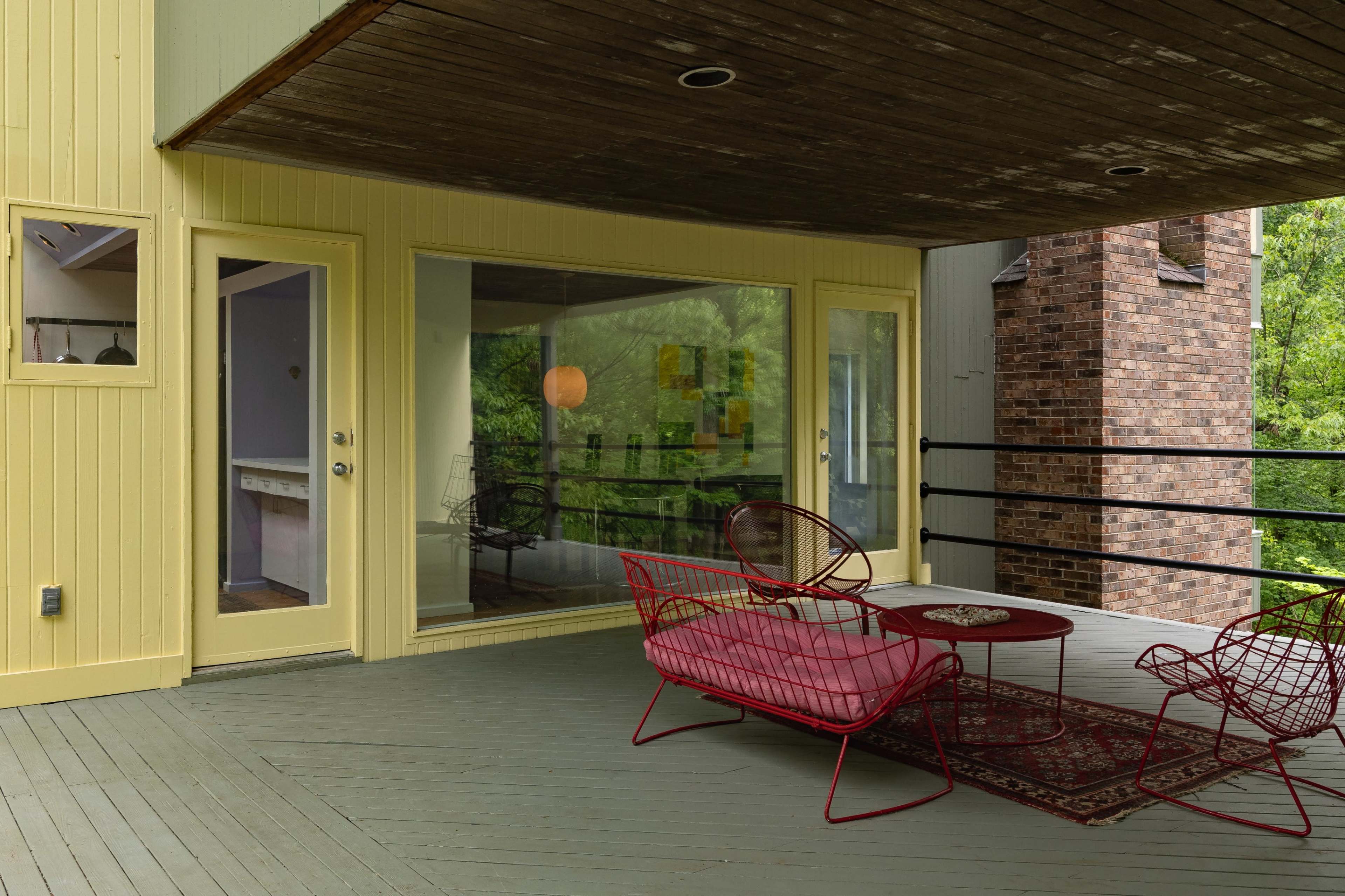 A modern, covered outdoor patio featuring two red wire chairs and a round table, with a view of greenery through large windows.