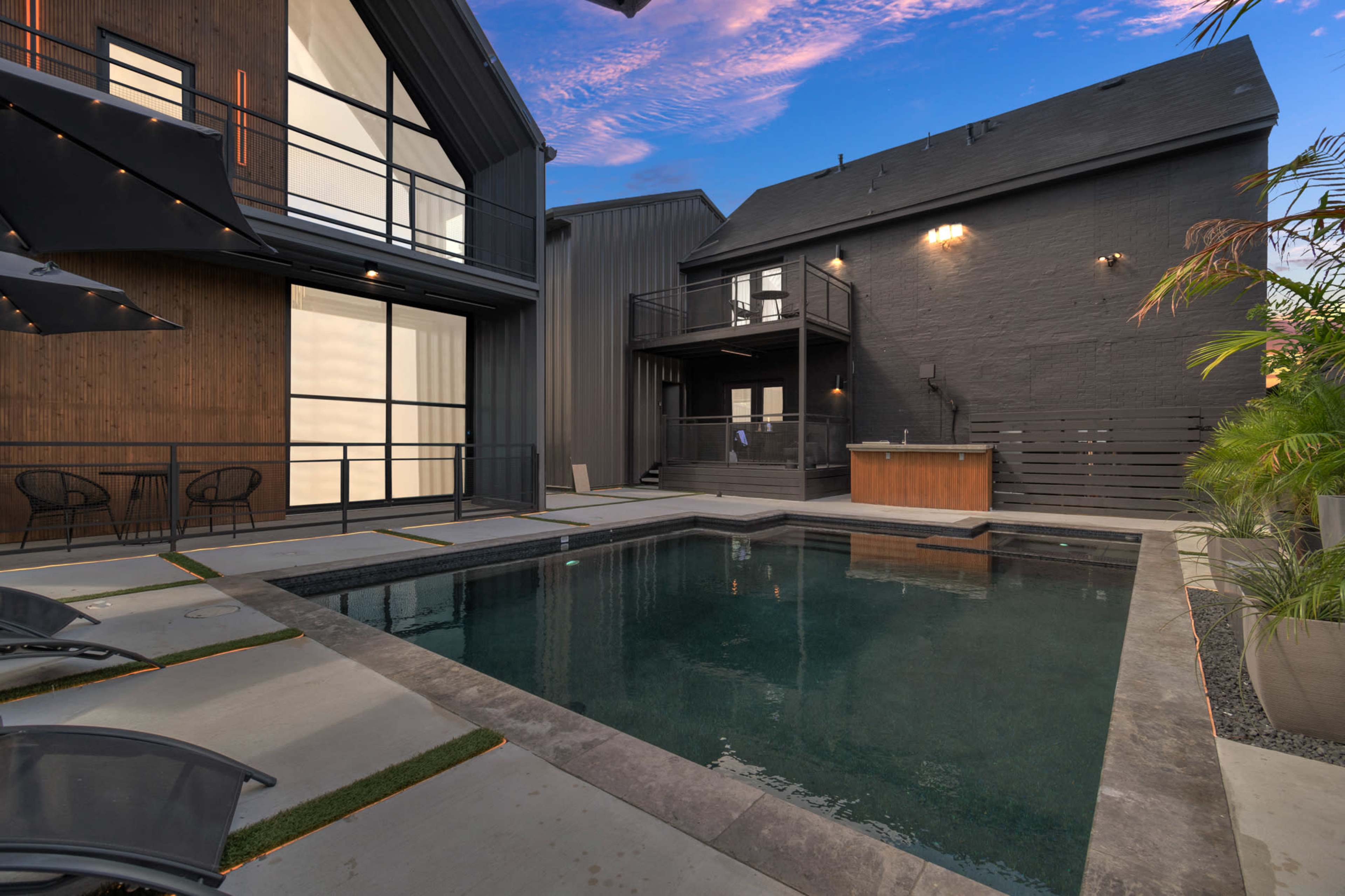 The image shows a modern backyard with a rectangular swimming pool surrounded by stone landscaping and two adjacent buildings, one featuring a wooden façade and the other a dark exterior.