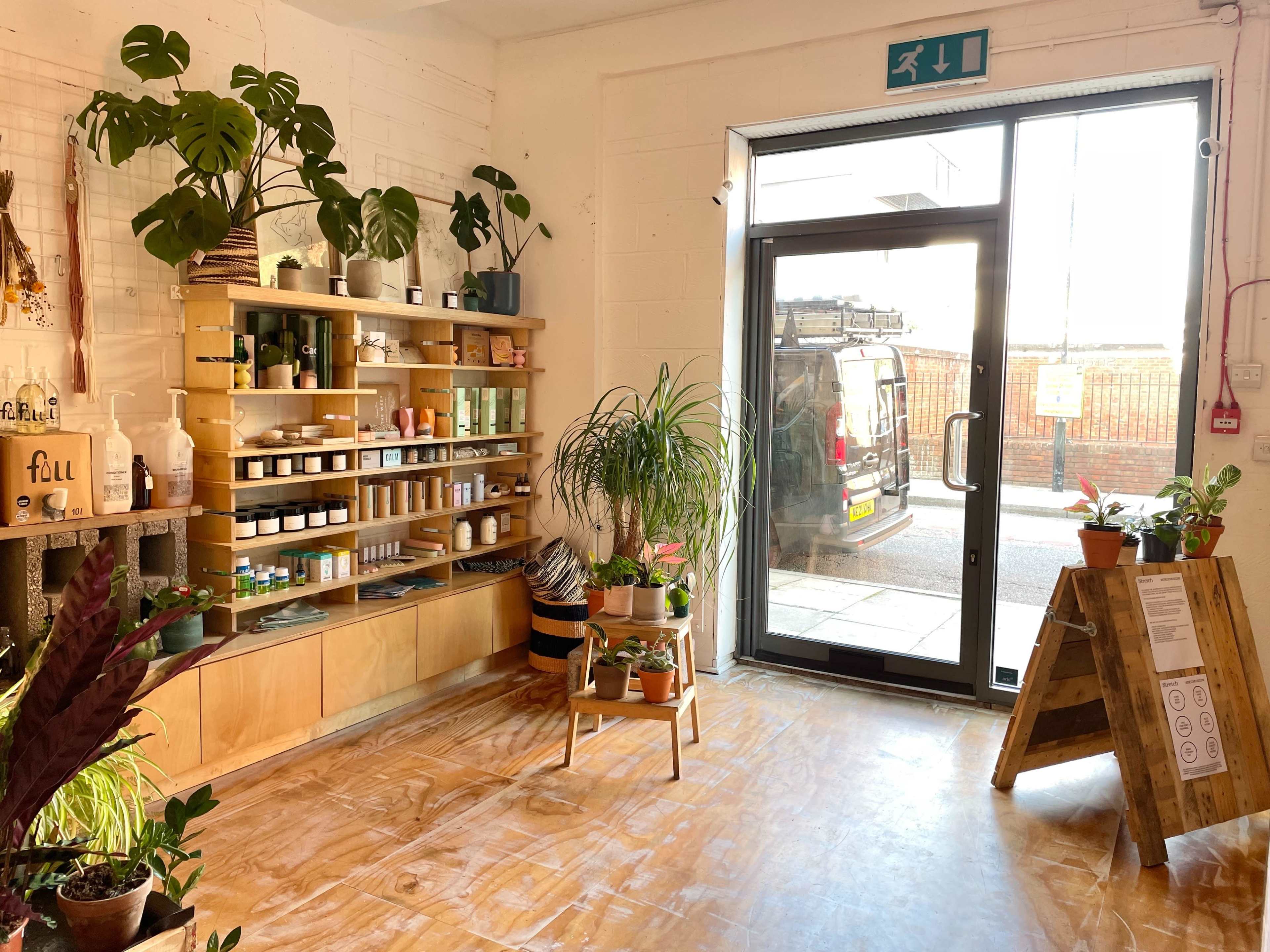 The image shows a bright, airy shop interior with wooden shelves stocked with various jars and plants, and a large glass door leading outside.