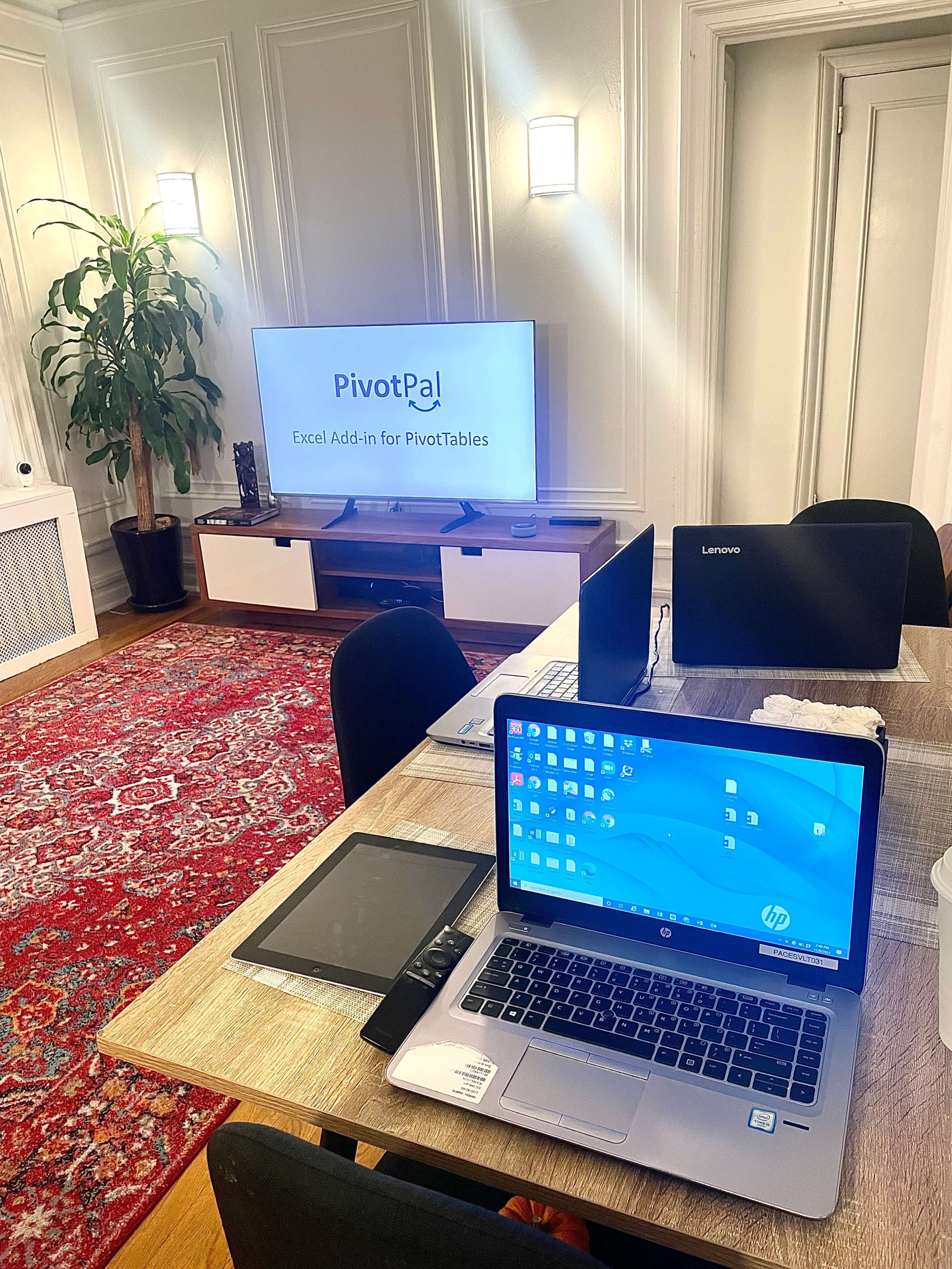 A laptop sits on a wooden table in a well-lit room with a large screen displaying "PivotPal Excel Add-in for PivotTables," while additional laptops and a plant are visible in the background.