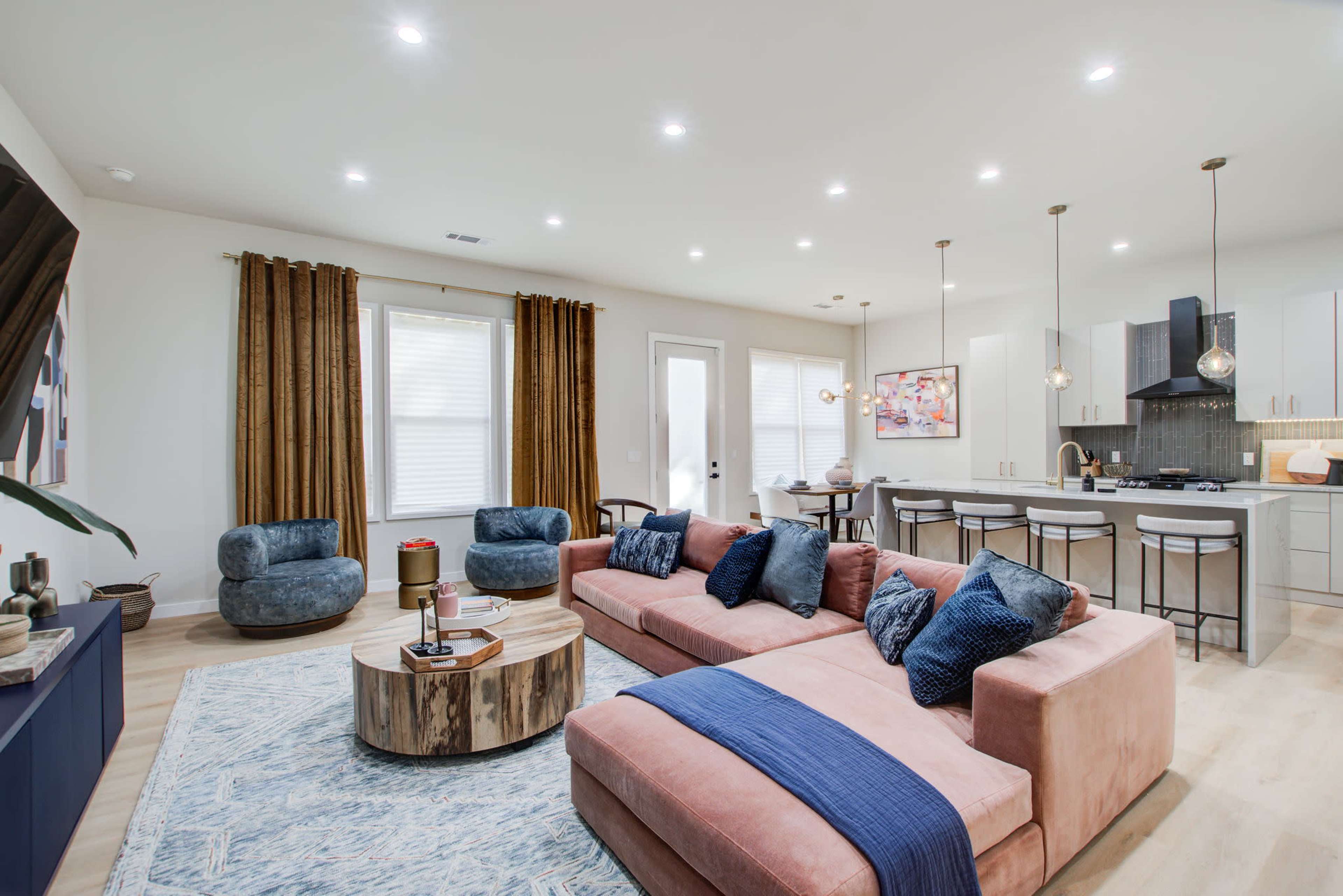A modern living room with a pink sectional sofa, blue accent chairs, a round wooden coffee table, and a kitchen area in the background featuring white cabinets and pendant lighting.