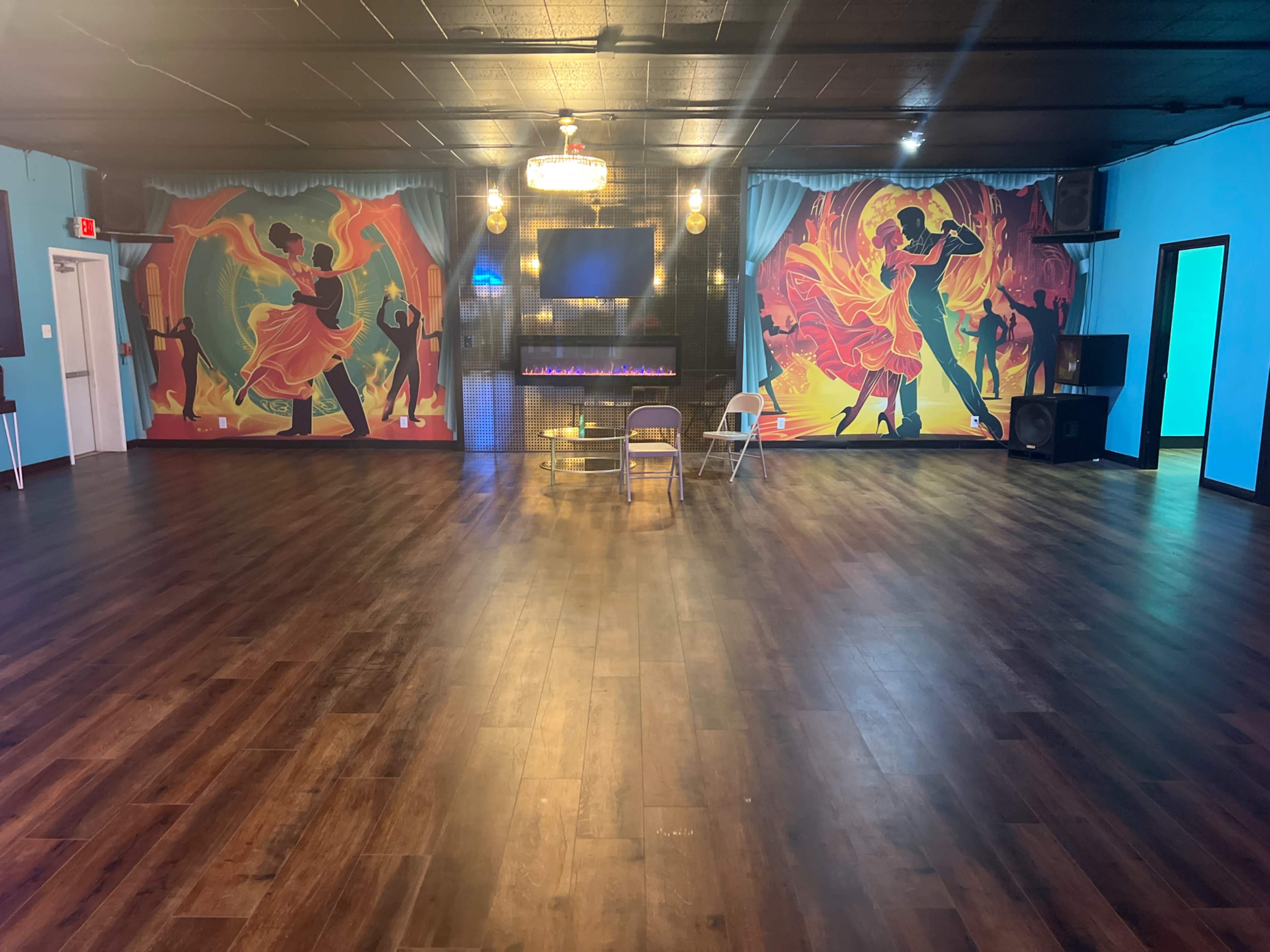 A spacious dance studio with colorful murals of dancers on the walls, a wooden floor, and a small seating area with a table and chairs in the center.