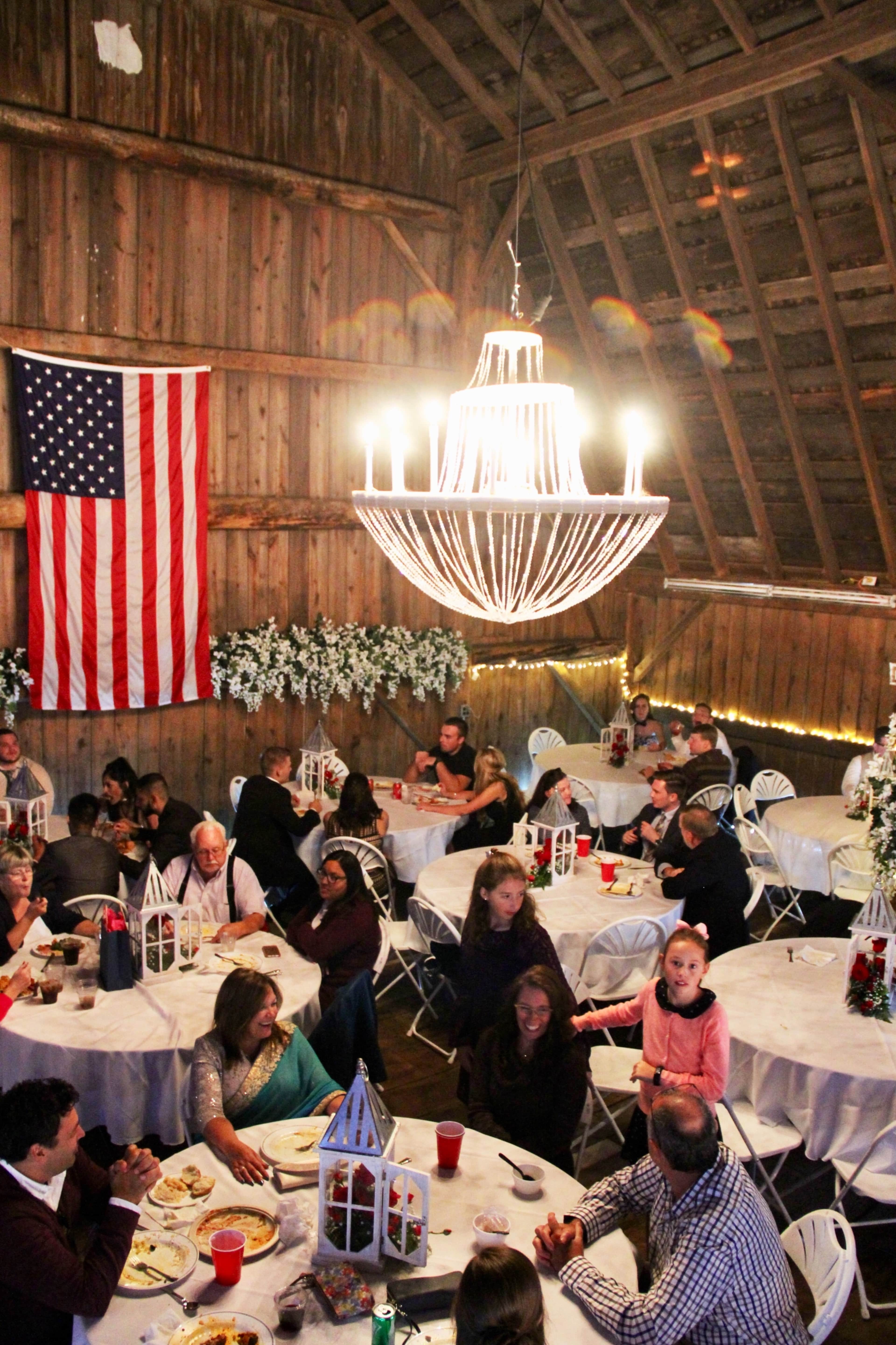 A well-lit barn interior features tables set for a gathering, with a large American flag hanging on the wall and guests enjoying food and conversation.