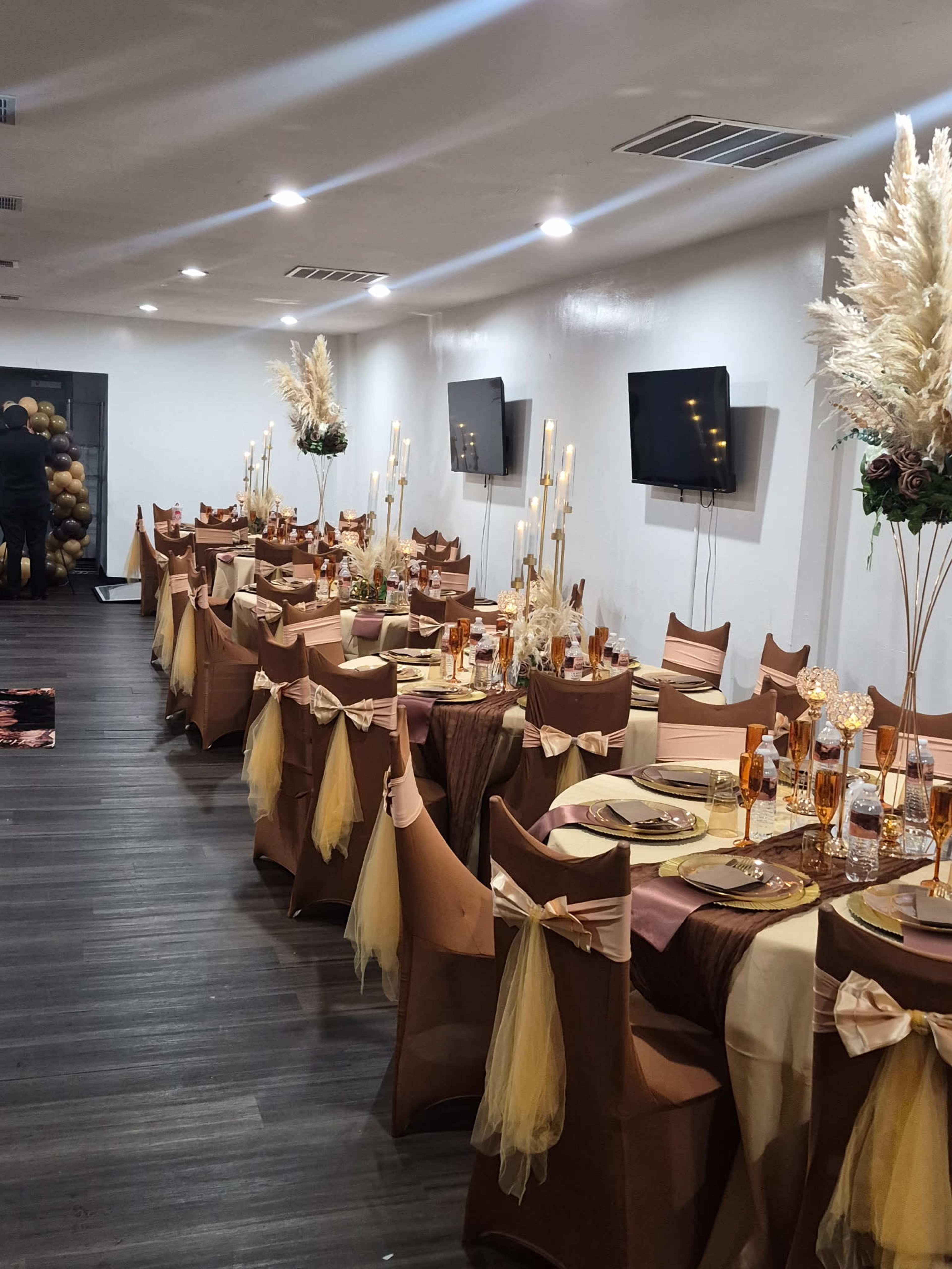 The image shows a elegantly decorated banquet hall with multiple tables set for an event, featuring gold and brown table settings, tall candle holders, and decorative floral arrangements.