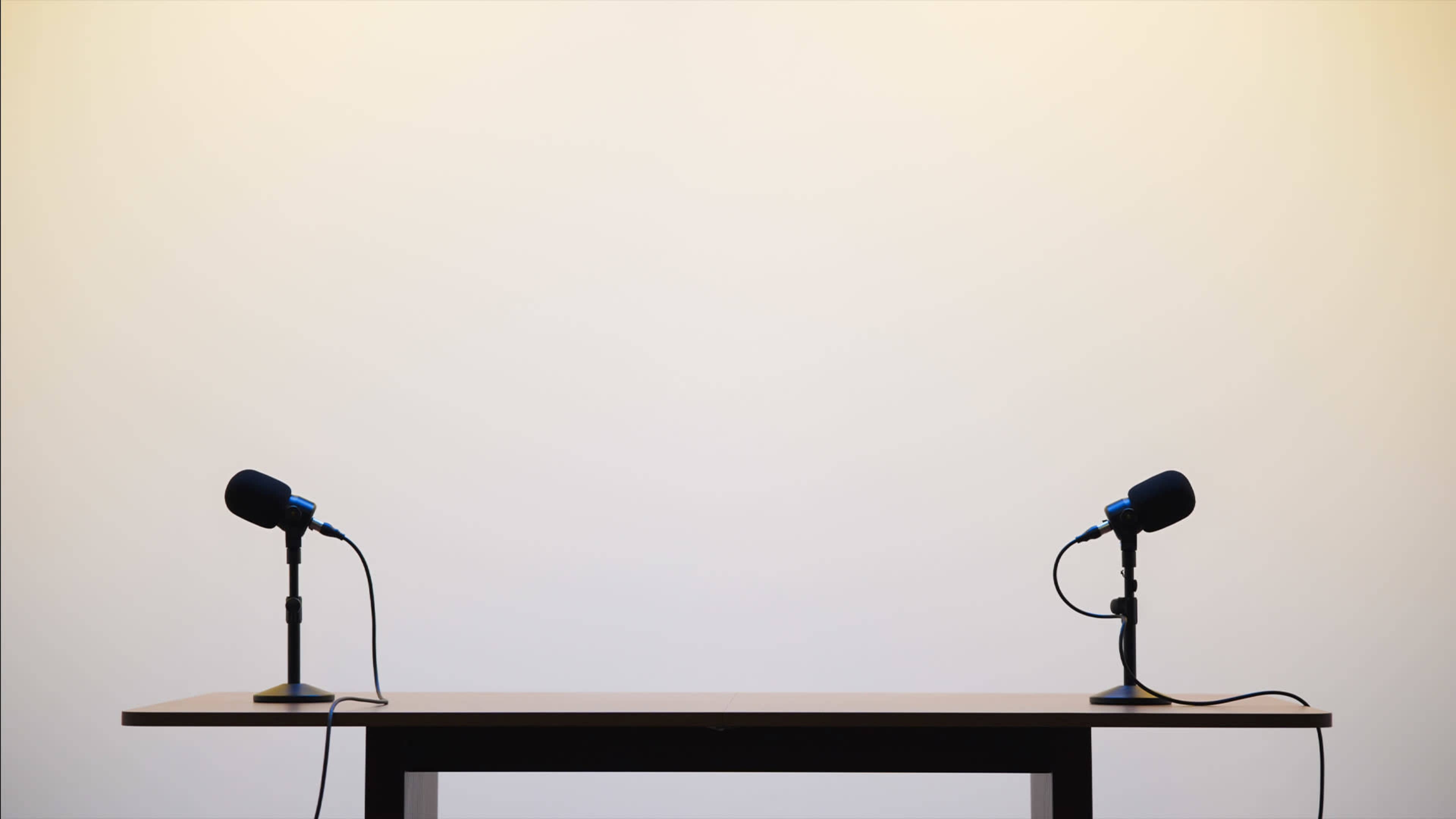 A simple wooden table sits center stage, flanked by two microphones on stands, against a plain light backdrop.