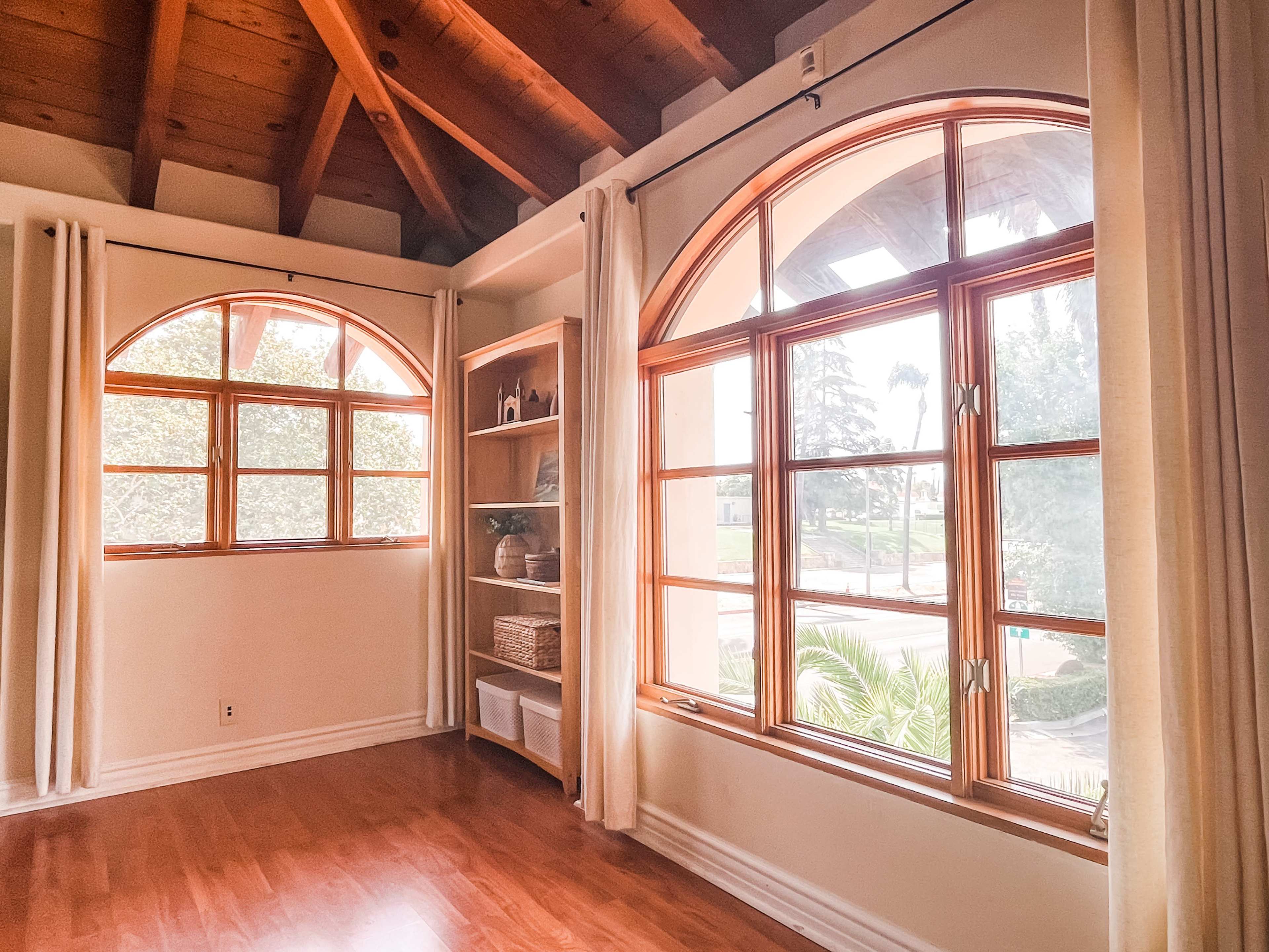 The image shows a spacious interior room with wooden beams and large arched windows overlooking a landscaped outdoor area.