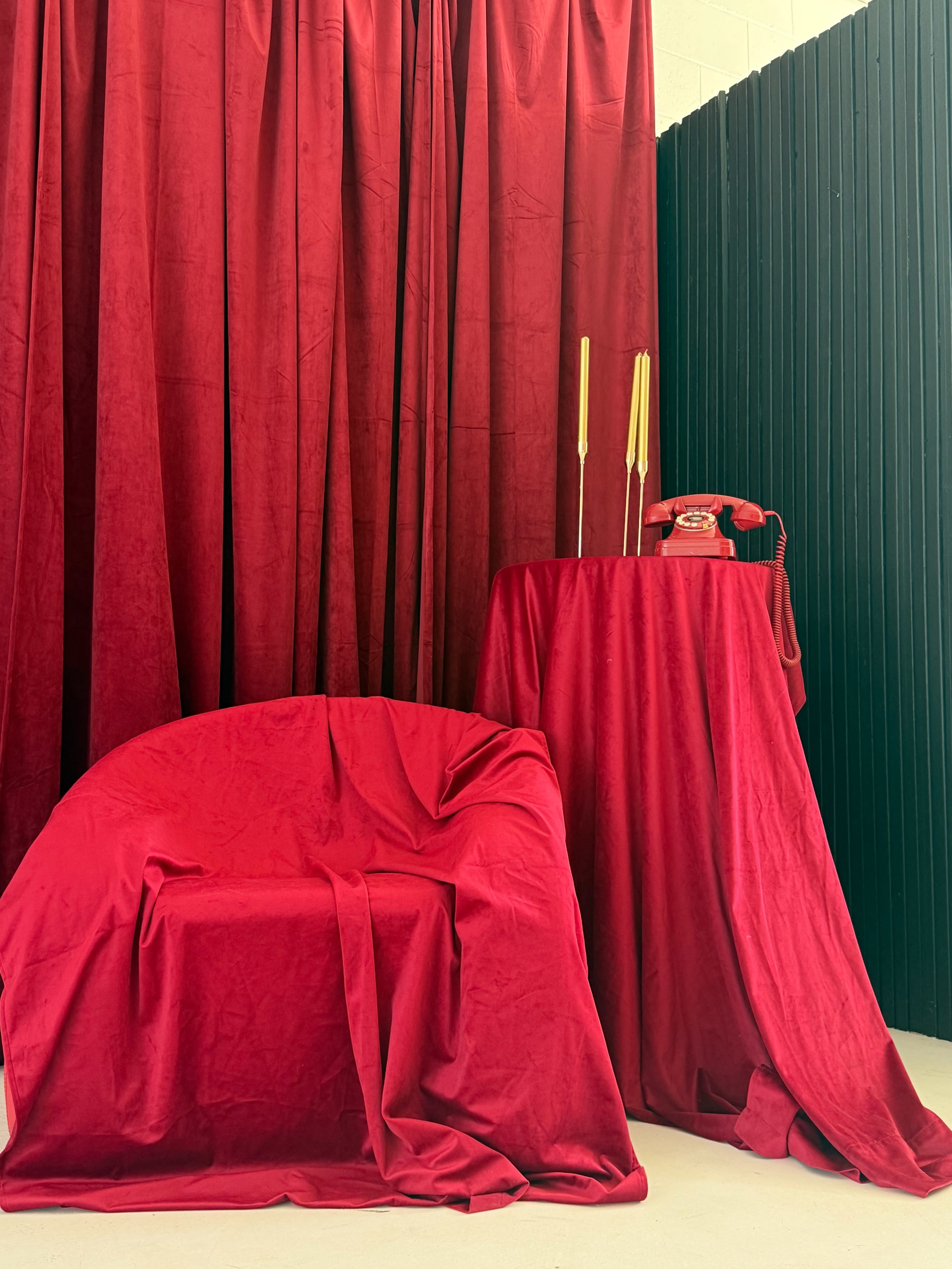 The scene features a red velvet chair draped with fabric next to a small table covered in red cloth, accompanied by two taper candles and a vintage red telephone against a backdrop of red curtains.
