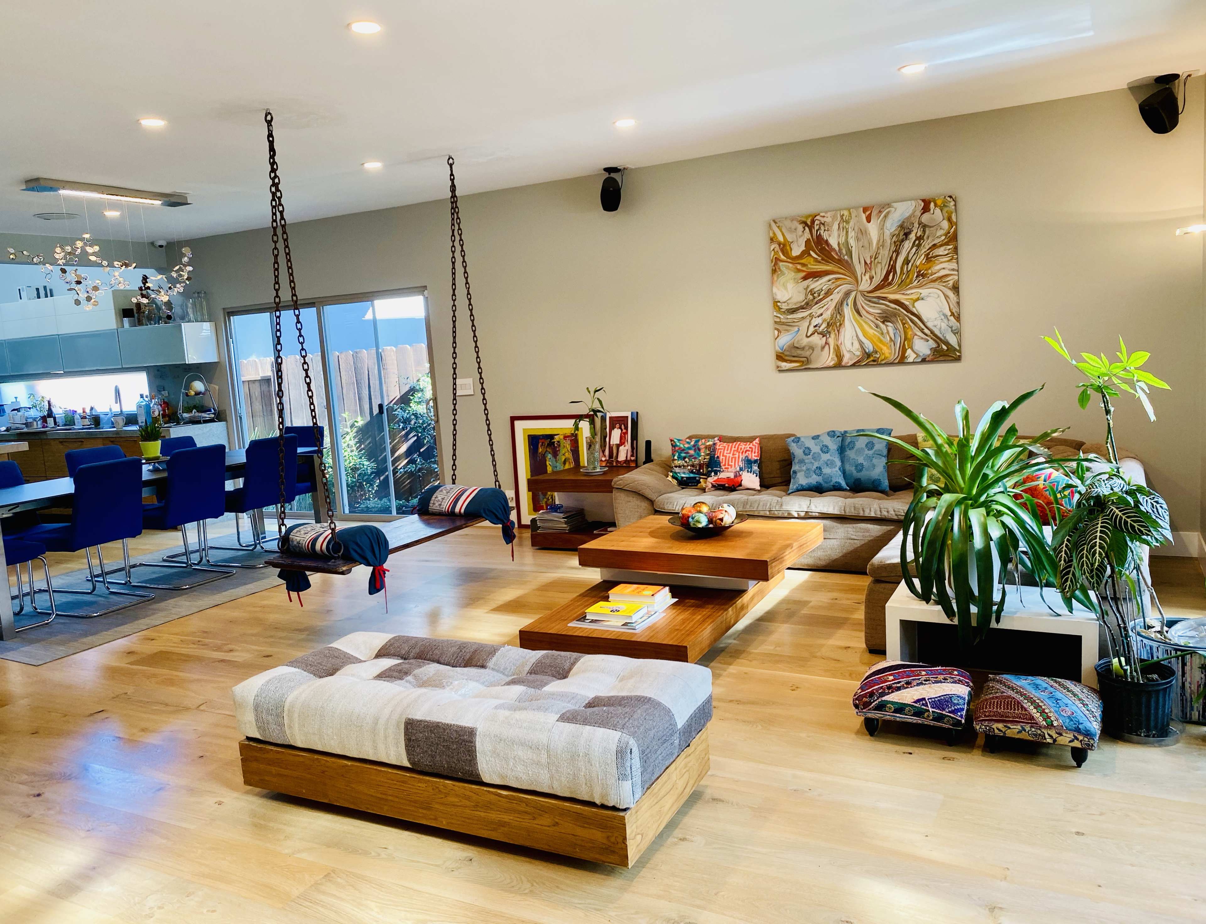 The image shows a modern living room with a wooden coffee table, a sectional sofa, two hanging chairs, and a dining area with blue barstools in the background.