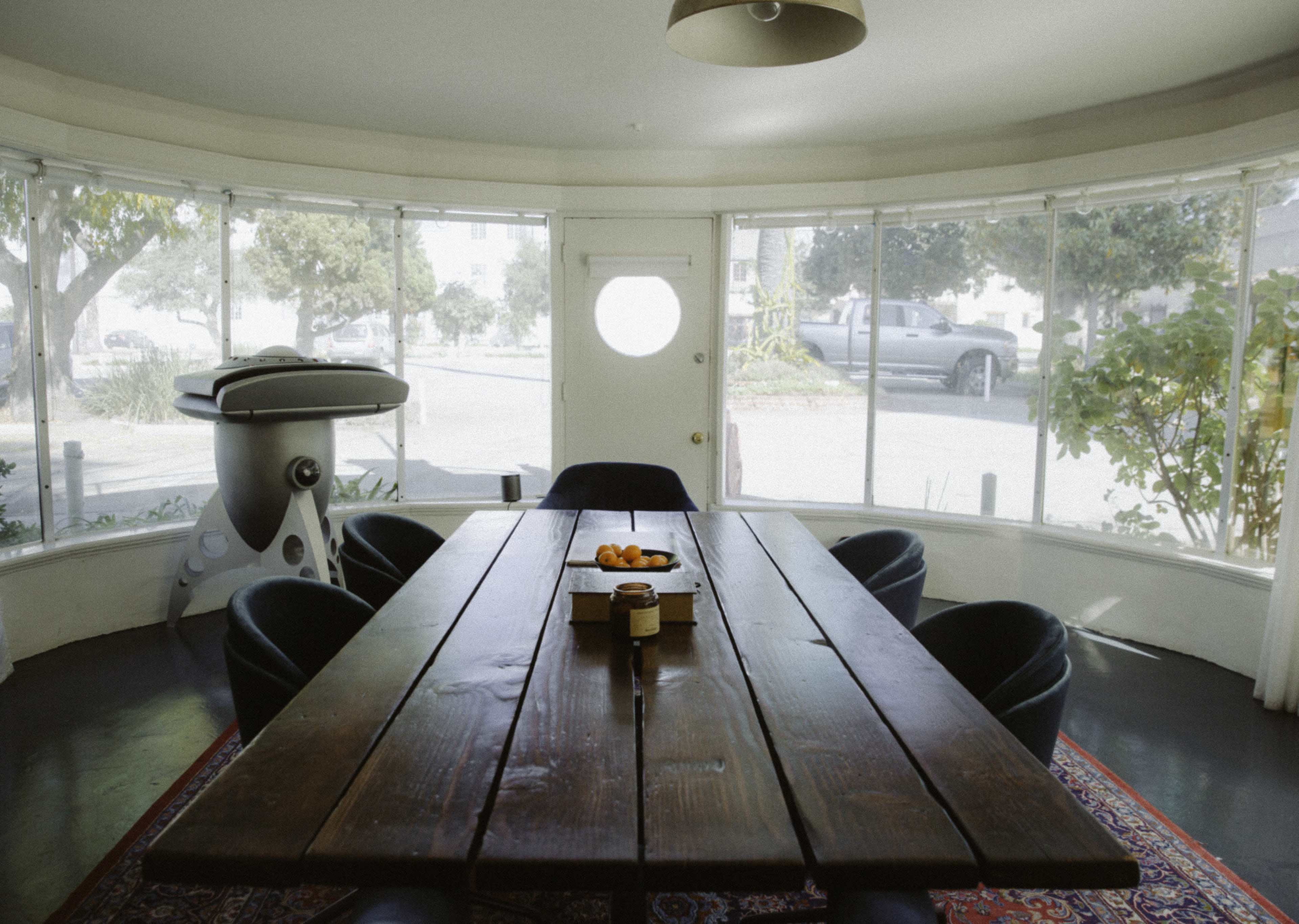 A large wooden dining table surrounded by black chairs is positioned in a bright, circular room with large windows and a door that opens to the exterior.