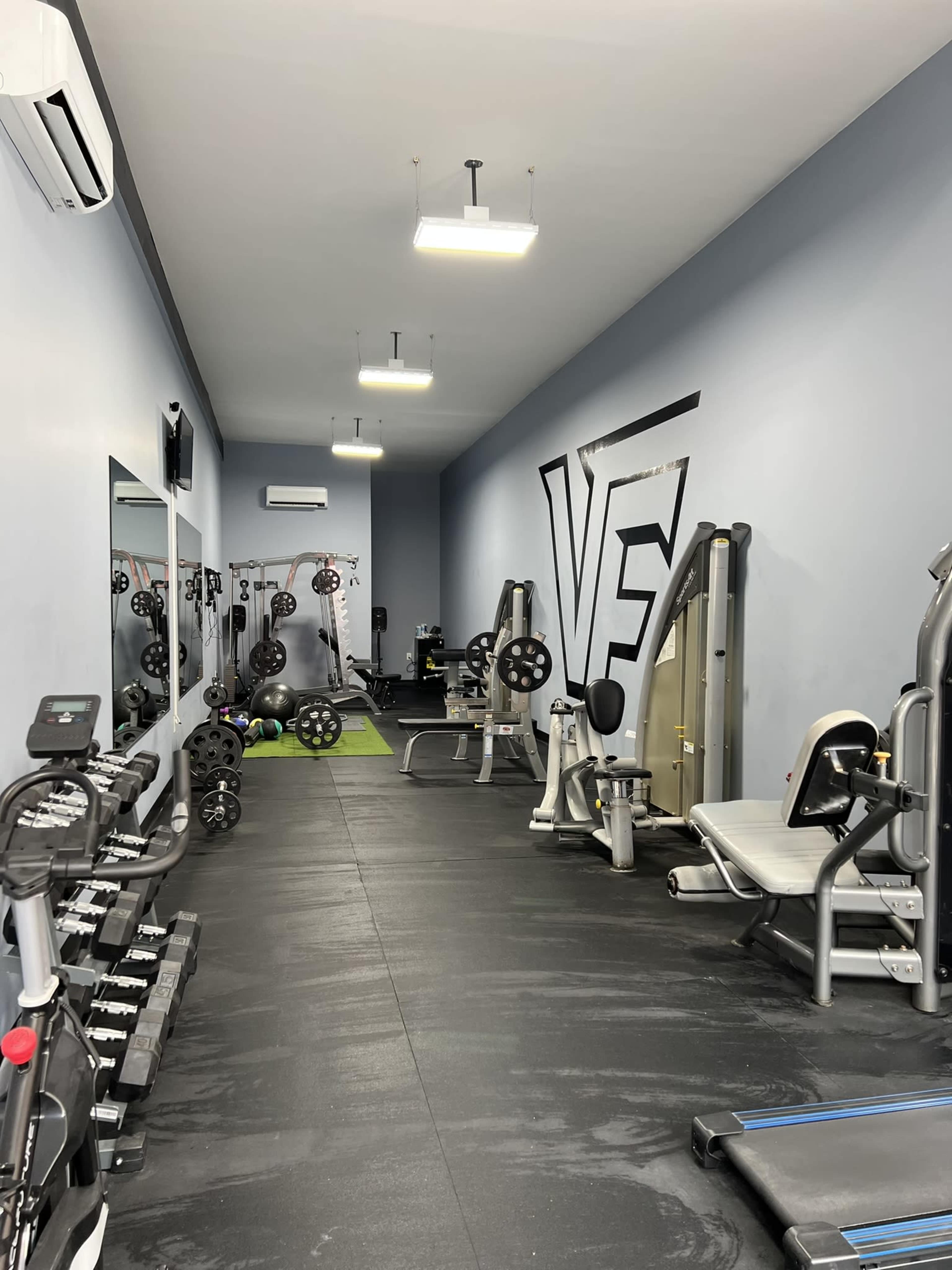 The image shows a gym interior featuring various weightlifting equipment, exercise machines, and free weights arranged along a wall.