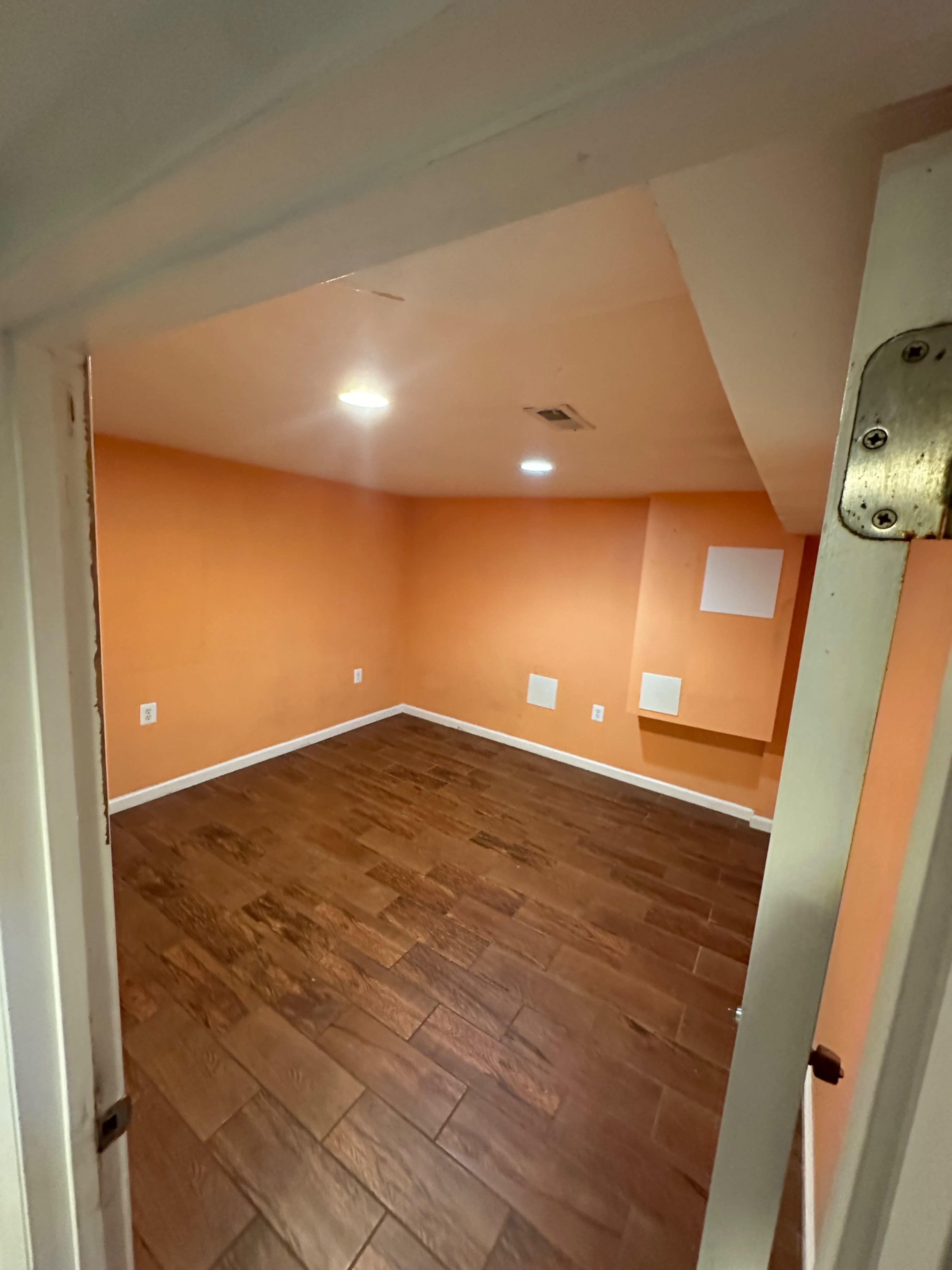 The image shows an empty room with orange walls, wooden flooring, and recessed lighting.