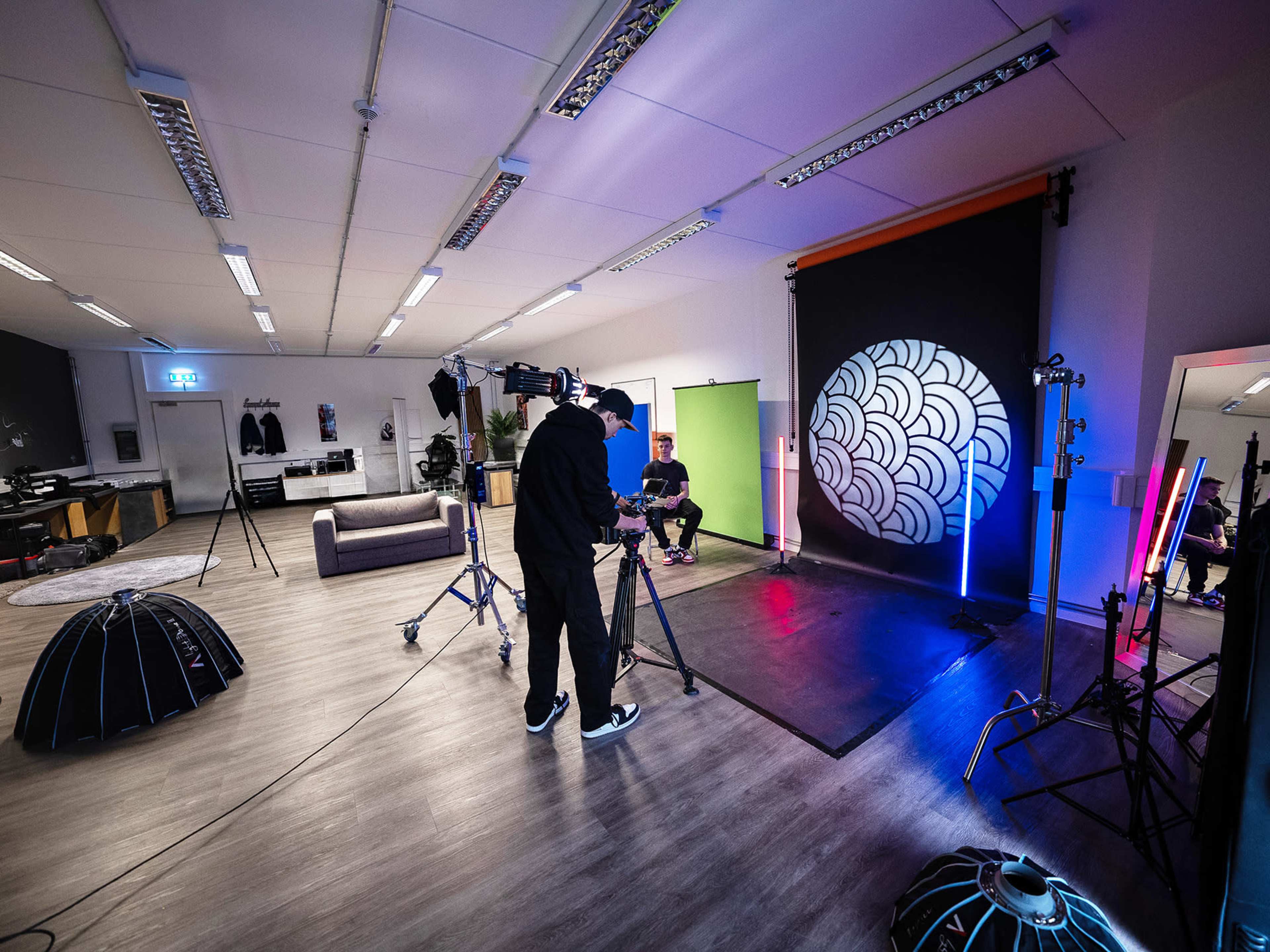 A photographer is setting up a camera in a studio filled with lighting equipment and a backdrop featuring a circular pattern.