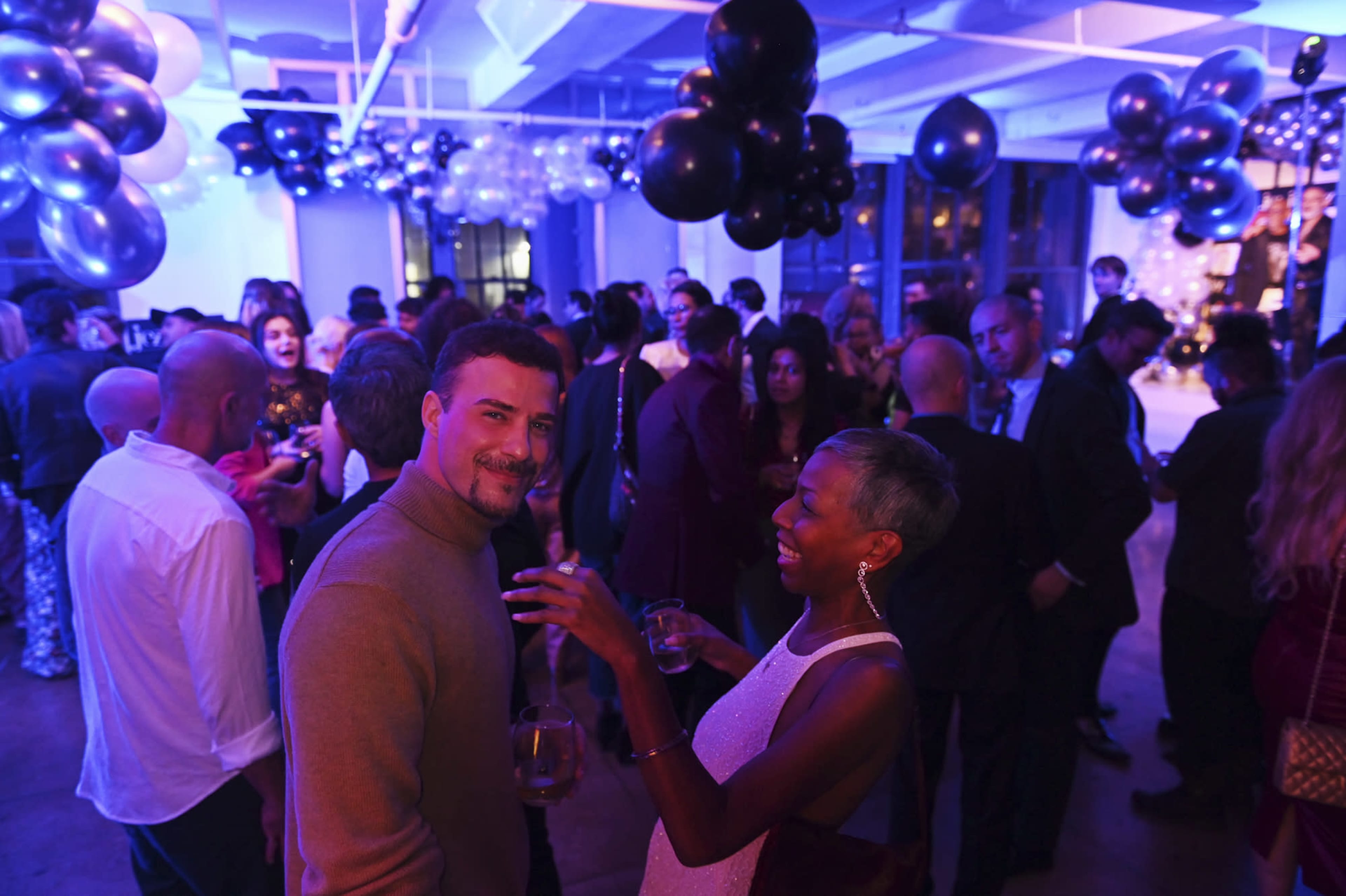 A crowd of people socializes at a party decorated with black and silver balloons under blue lighting.
