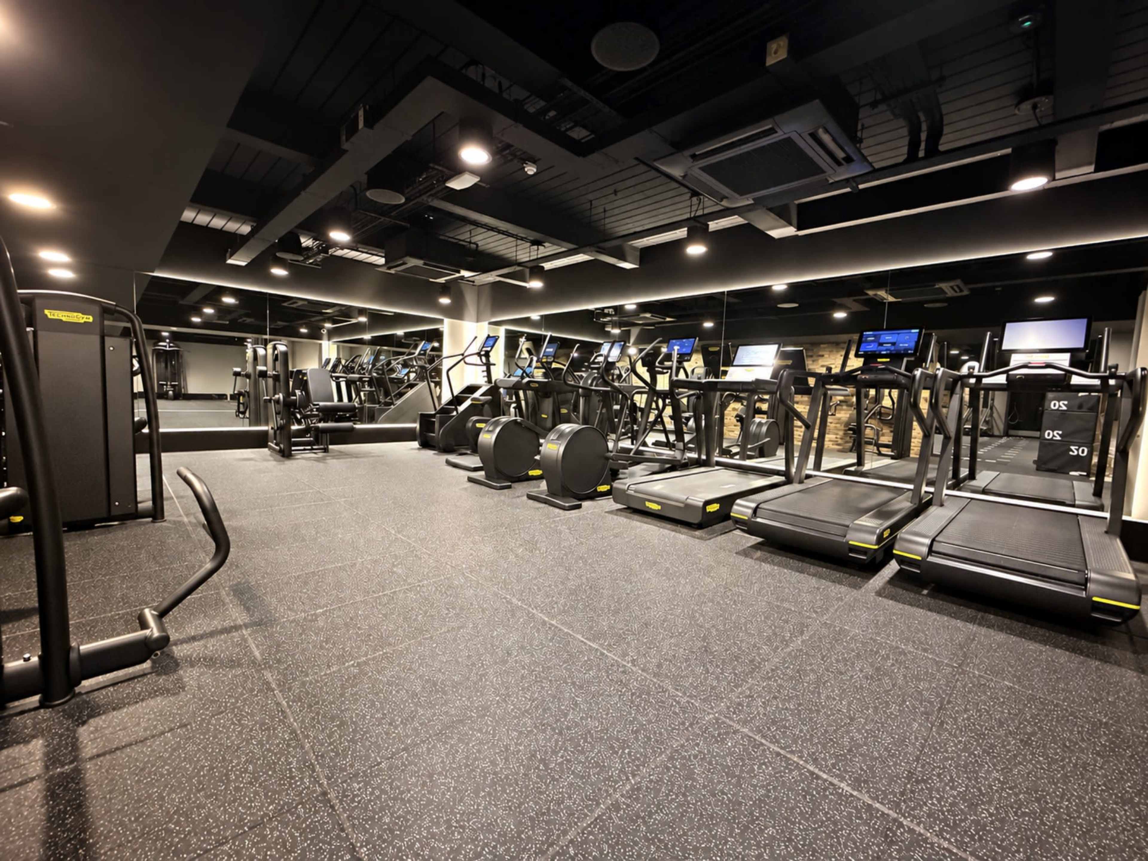 The image shows a modern gym equipped with various fitness machines, including treadmills, weight racks, and exercise bikes, under bright overhead lighting.
