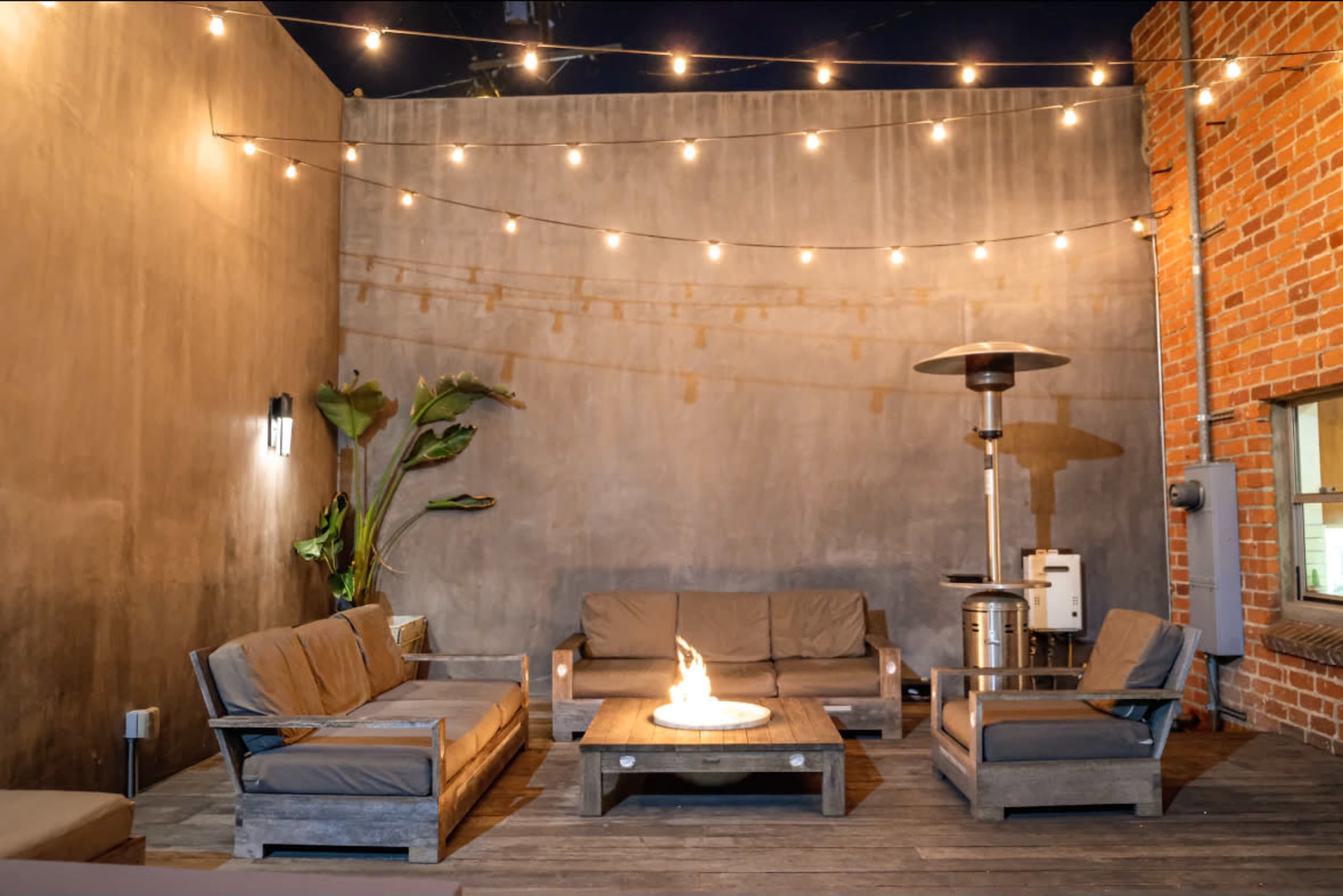 A cozy outdoor lounge area features seating arrangements around a central fire pit, illuminated by string lights against a textured wall.