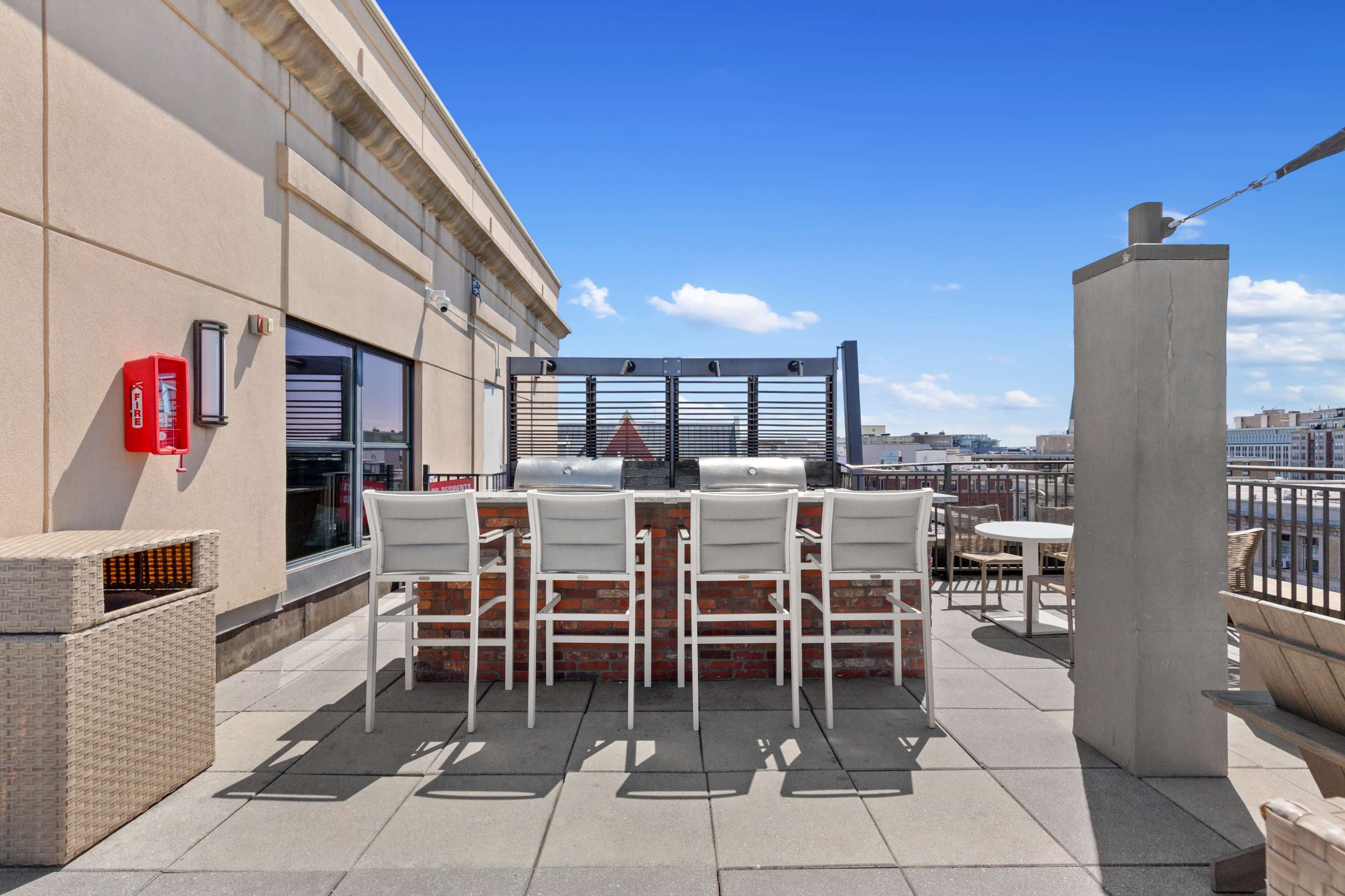 The image shows a rooftop terrace with a barbecue grill and several white barstools arranged around a brick counter, along with a clear blue sky in the background.